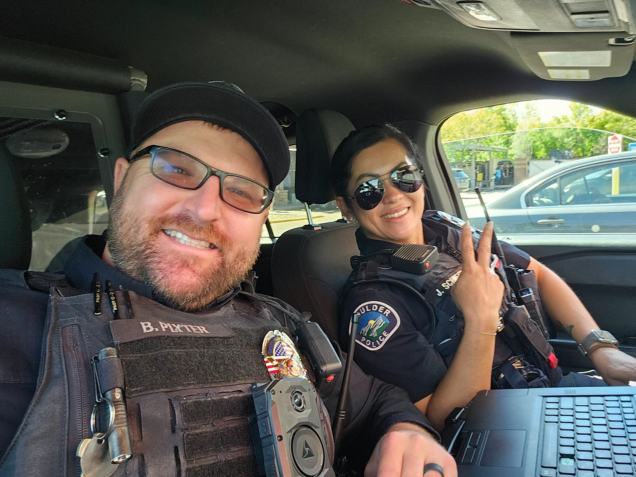 Two uniformed police officers smile for a selfie from the front seats of a patrol car.