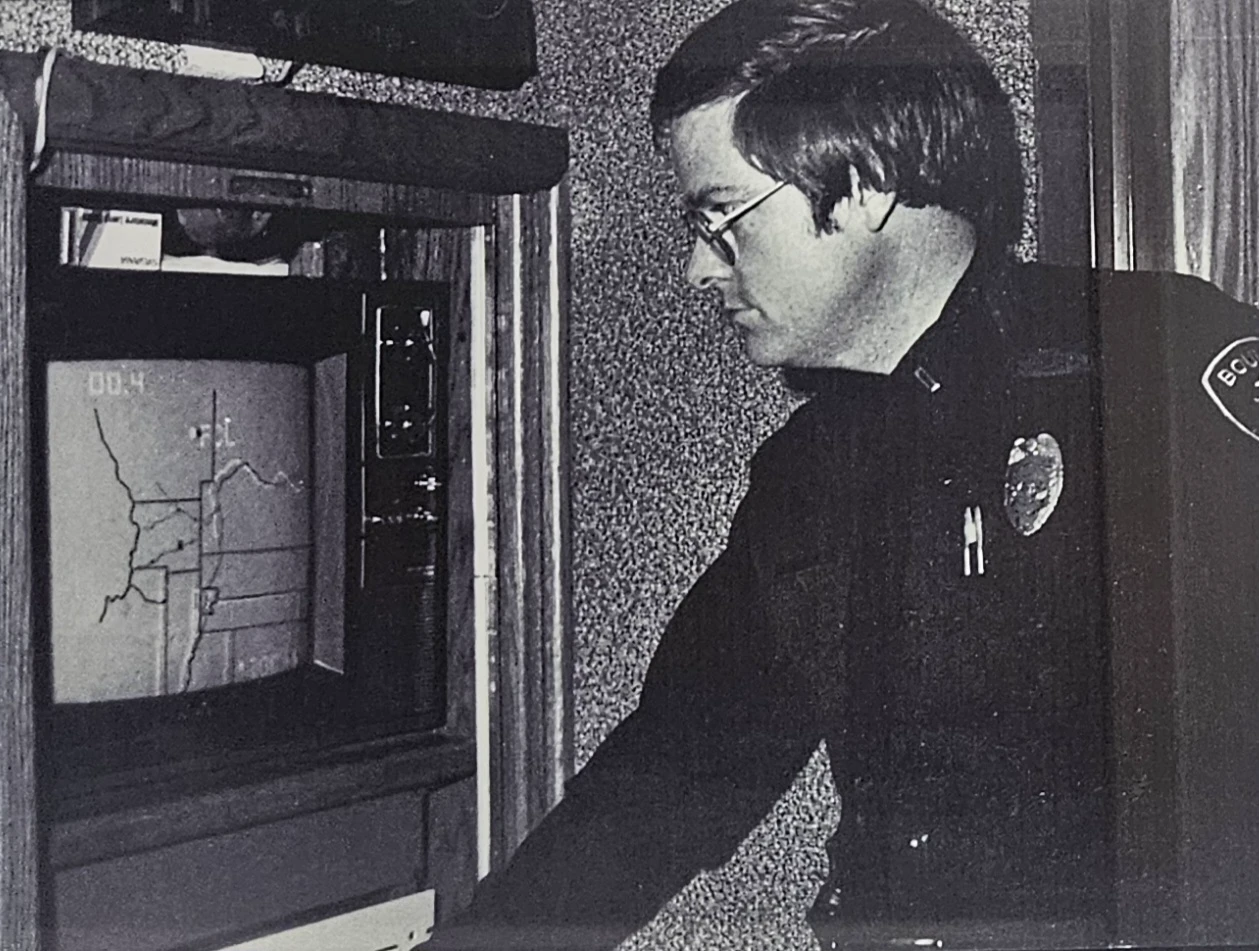 Historical black-and-white photo of a Boulder police officer studying a map displayed on an early computer monitor.