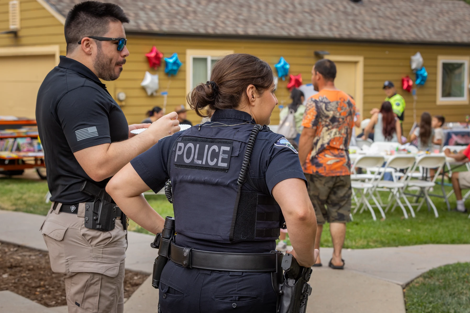 A uniformed police officer and a man in a black polo shirt stand at the edge of an outdoor gathering with balloons, tables, and chairs.