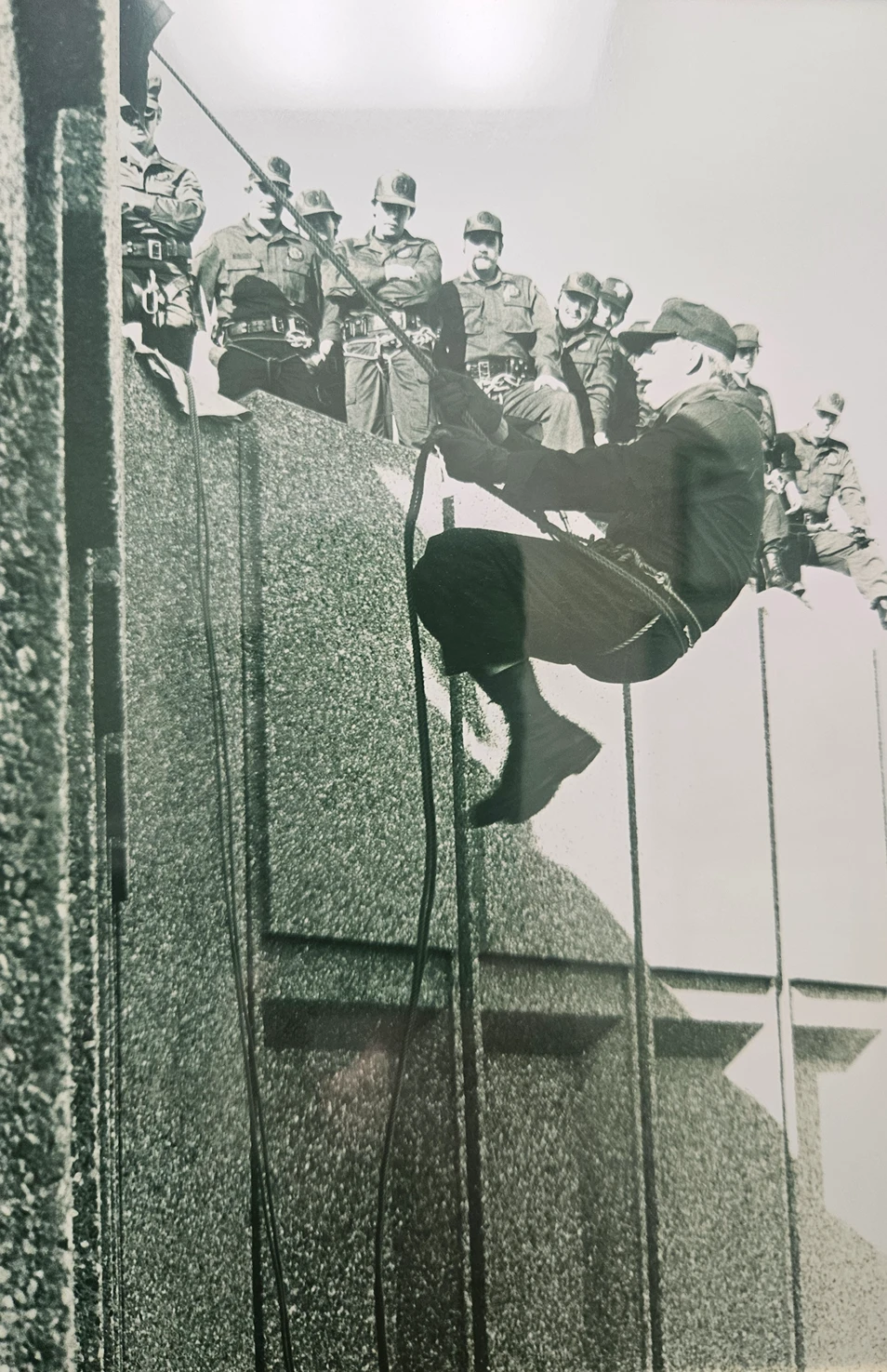 Historical black-and-white photo of a person rappelling down the side of a building as uniformed officers watch from the rooftop.