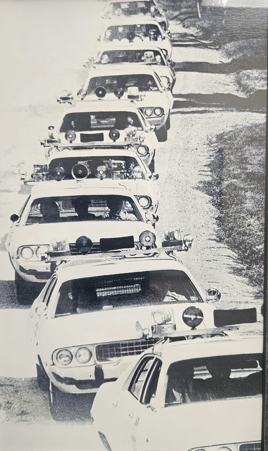 Historical black-and-white photo of a long line of Boulder police patrol cars with roof lights driving in procession down a rural road.