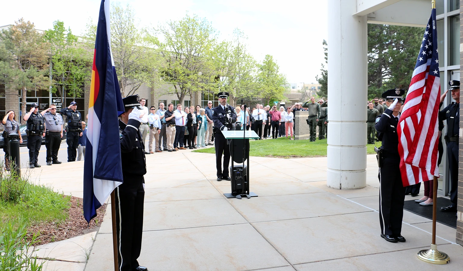 Officers and community members gather at an outdoor ceremony with the Colorado and American flags.