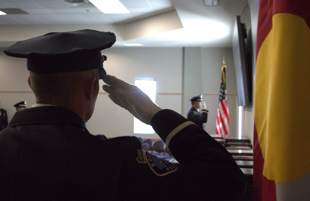 An honor guard officer salutes beside the Colorado state flag at a ceremony.