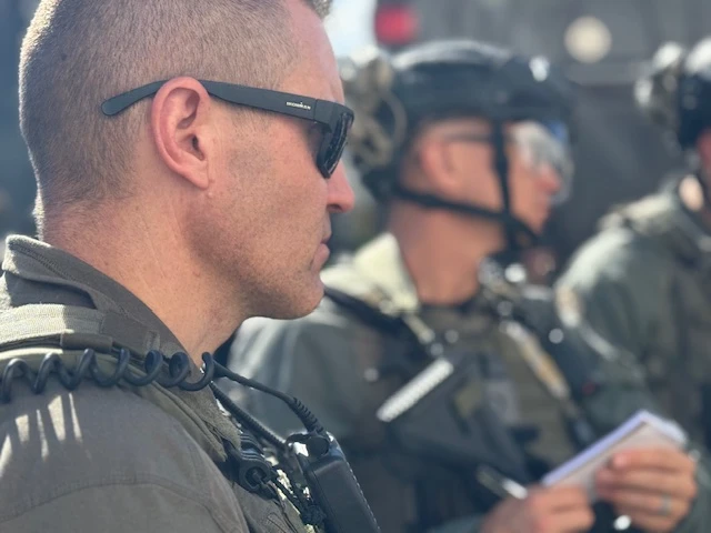 Officers in tactical gear and helmets stand together outdoors, with one officer in sunglasses in the foreground.