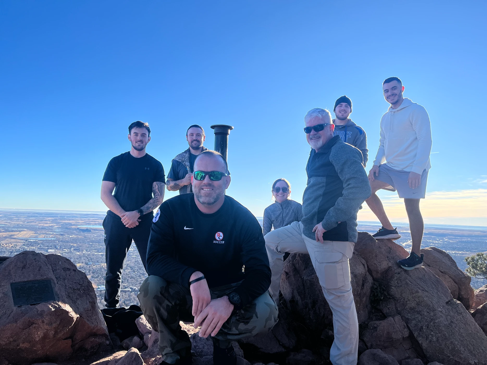 A group of seven people pose on red rocks at a mountain summit under a clear blue sky, with a wide plain visible below.
