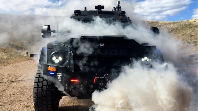 An armored police vehicle drives toward the camera through a cloud of dust on a dirt road.