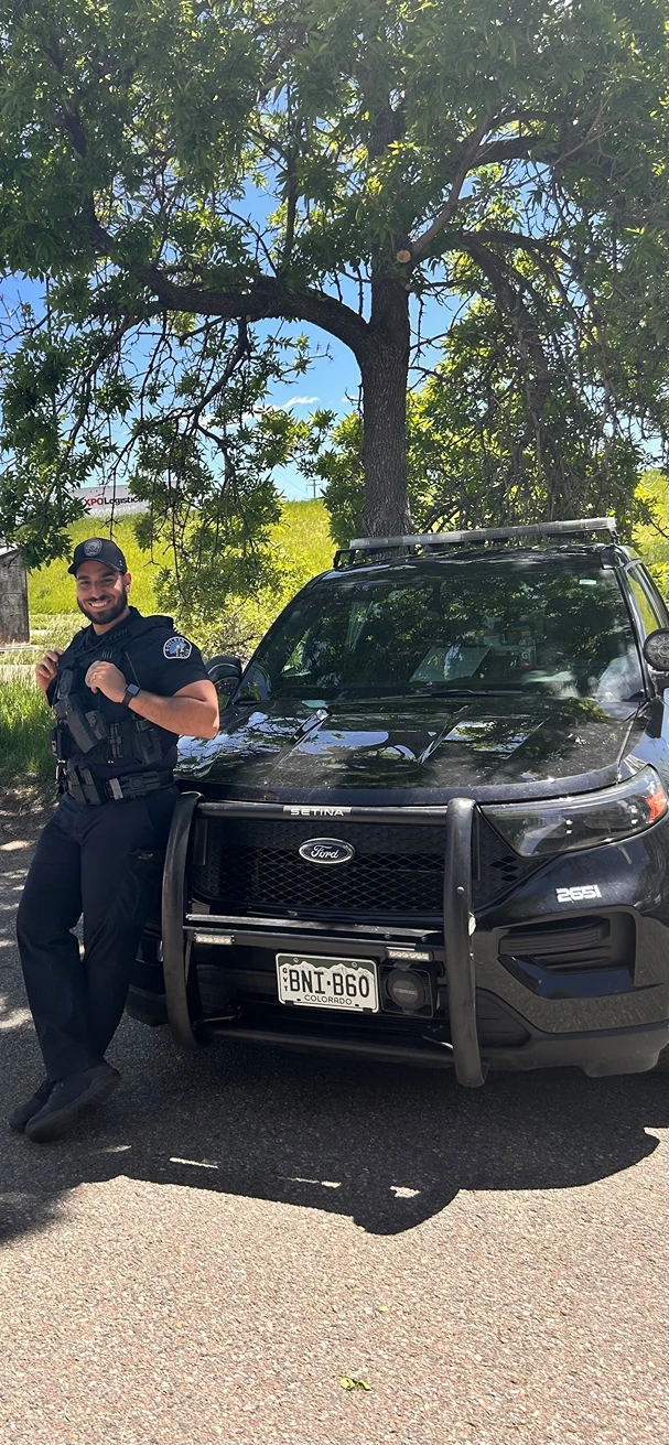 A smiling police officer in uniform leans against the front of a black police SUV parked under a large tree on a sunny day.