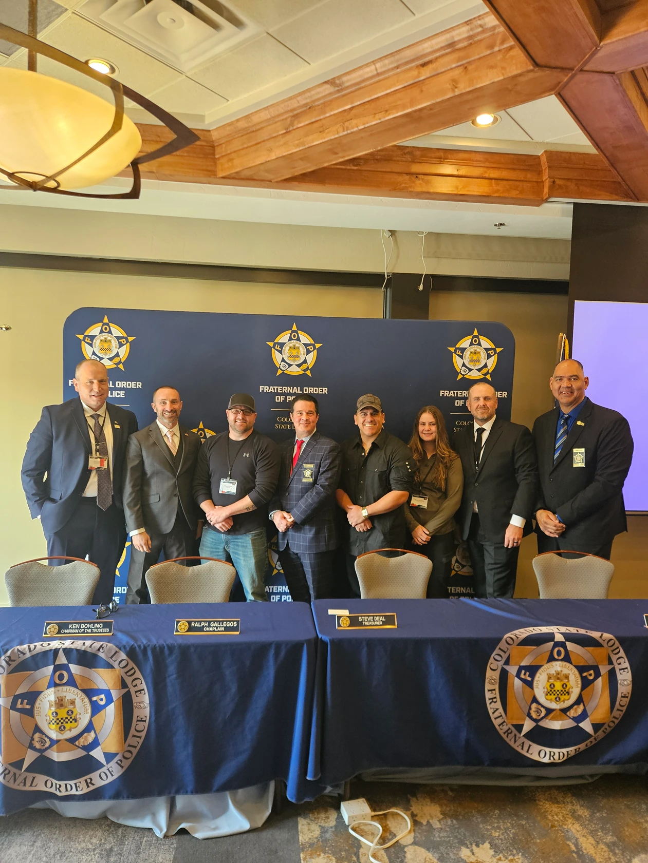 Eight people pose in front of a Fraternal Order of Police backdrop, standing behind tables draped with Fraternal Order of Police tablecloths.