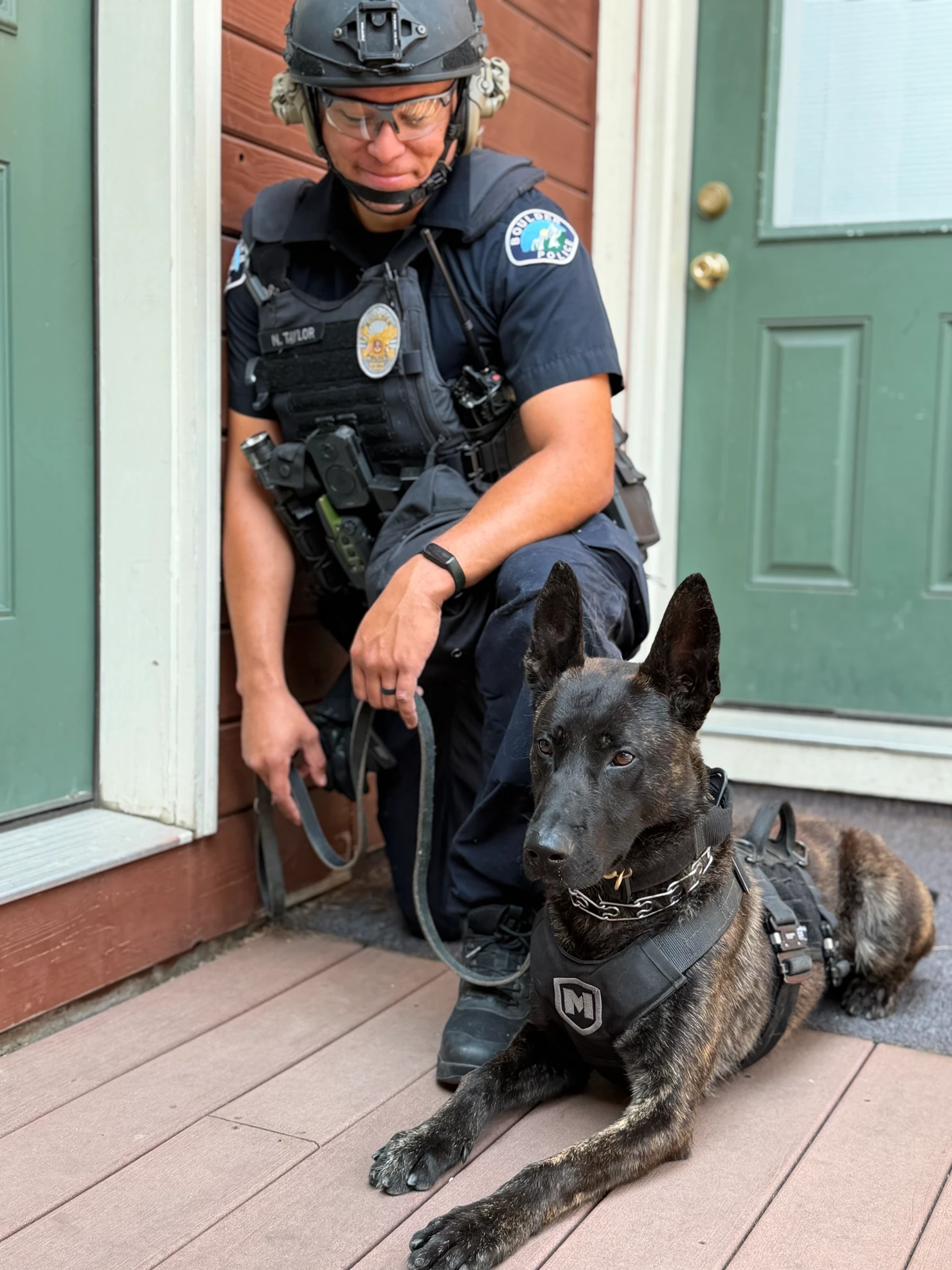 A police officer in a helmet and tactical vest kneels on a wooden porch holding the leash of a K9 dog lying beside them.
