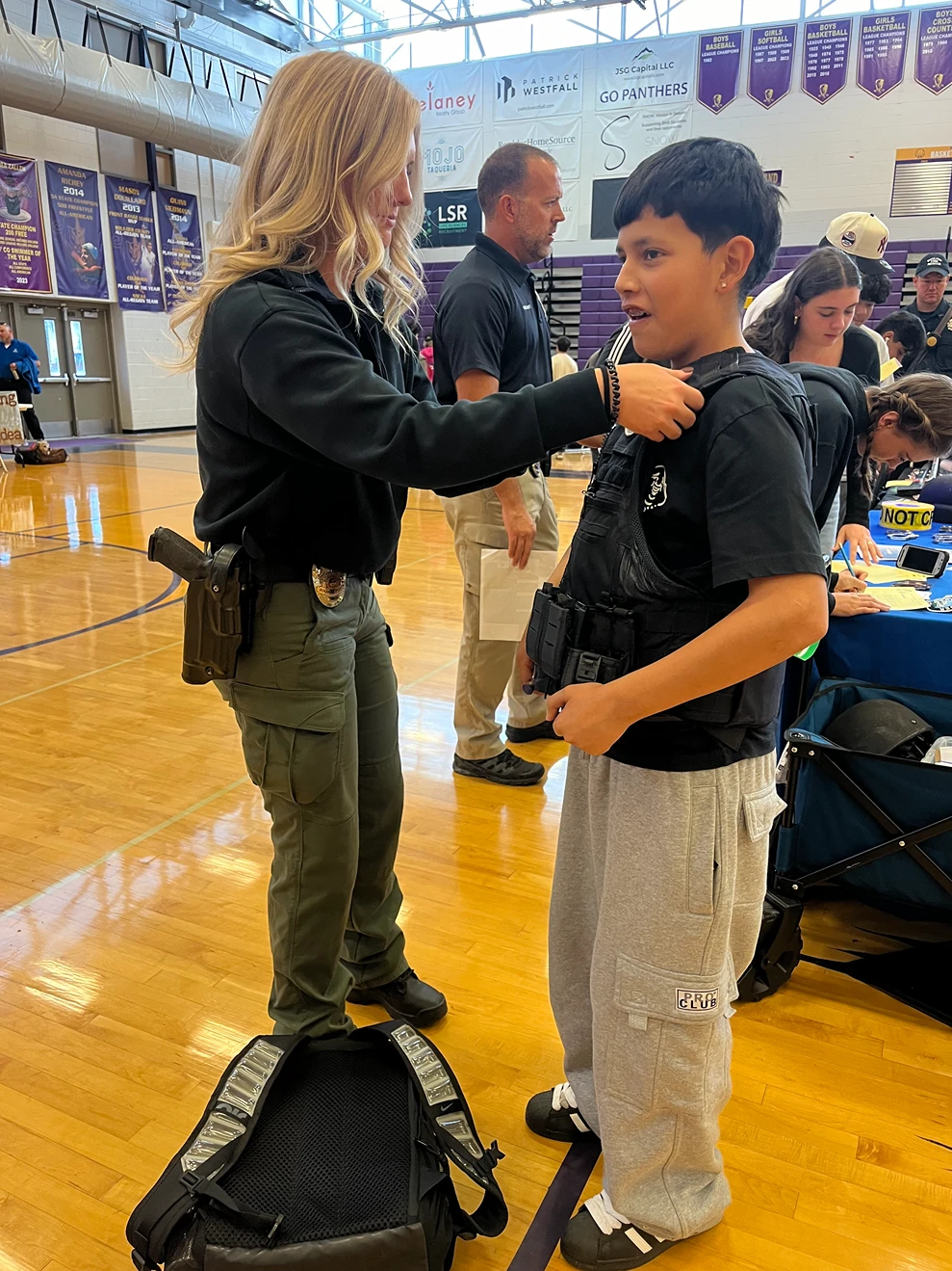 An officer helps a young person try on a tactical vest at a community event in a school gymnasium.