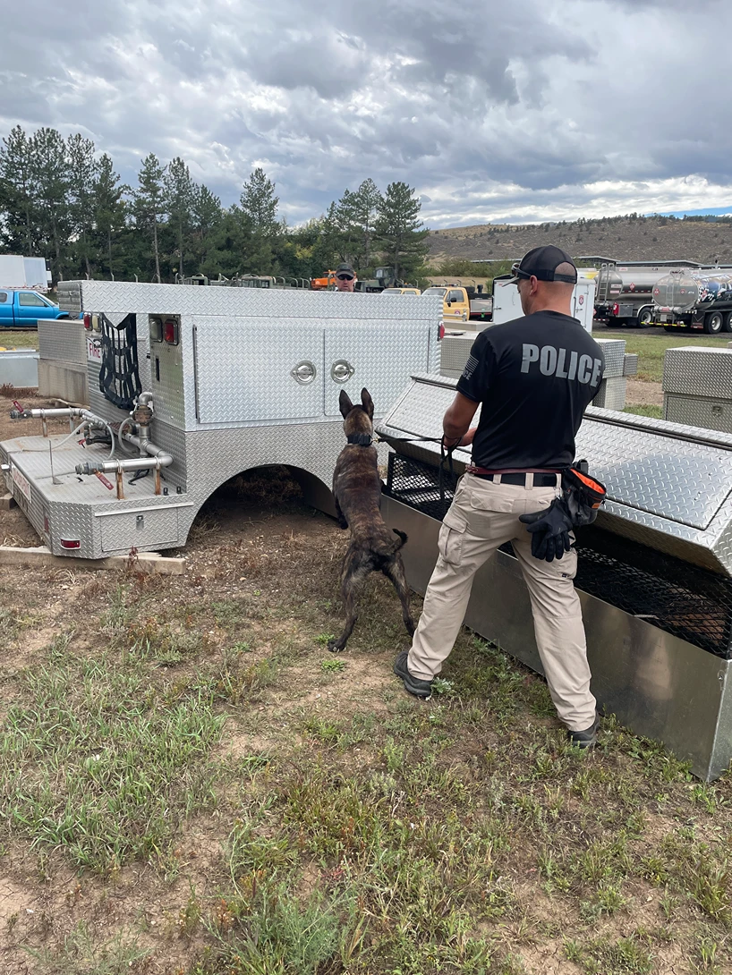 An officer in a police shirt leads a K9 dog on a leash past metal truck utility boxes at an outdoor site.