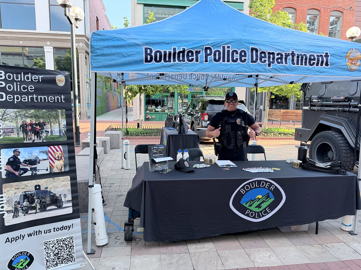 An officer stands behind a Boulder Police Department booth with a blue canopy and branded table at an outdoor community event, beside a recruiting banner.