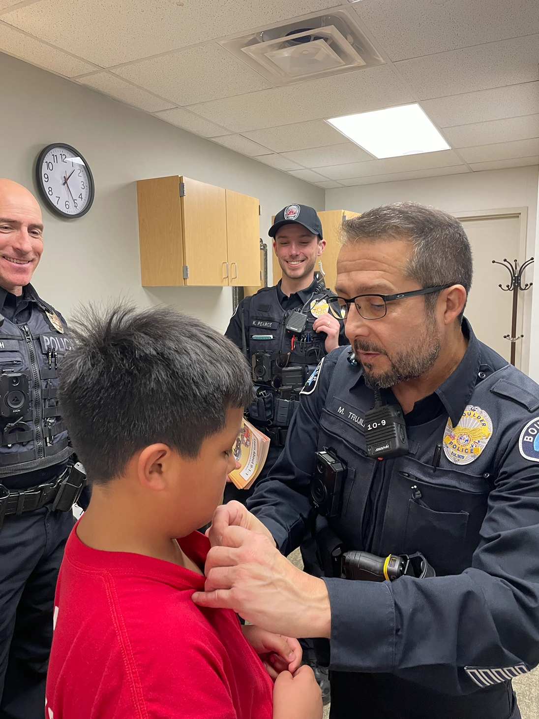 A police officer pins an item onto a boy's red shirt while two other uniformed officers look on, smiling.