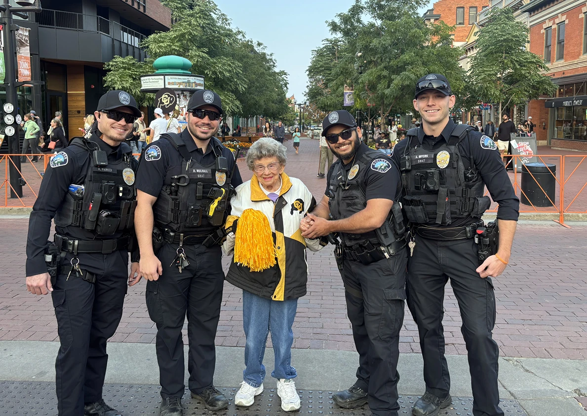 Four uniformed police officers pose with a smiling older woman holding yellow pom-poms on a brick pedestrian street.
