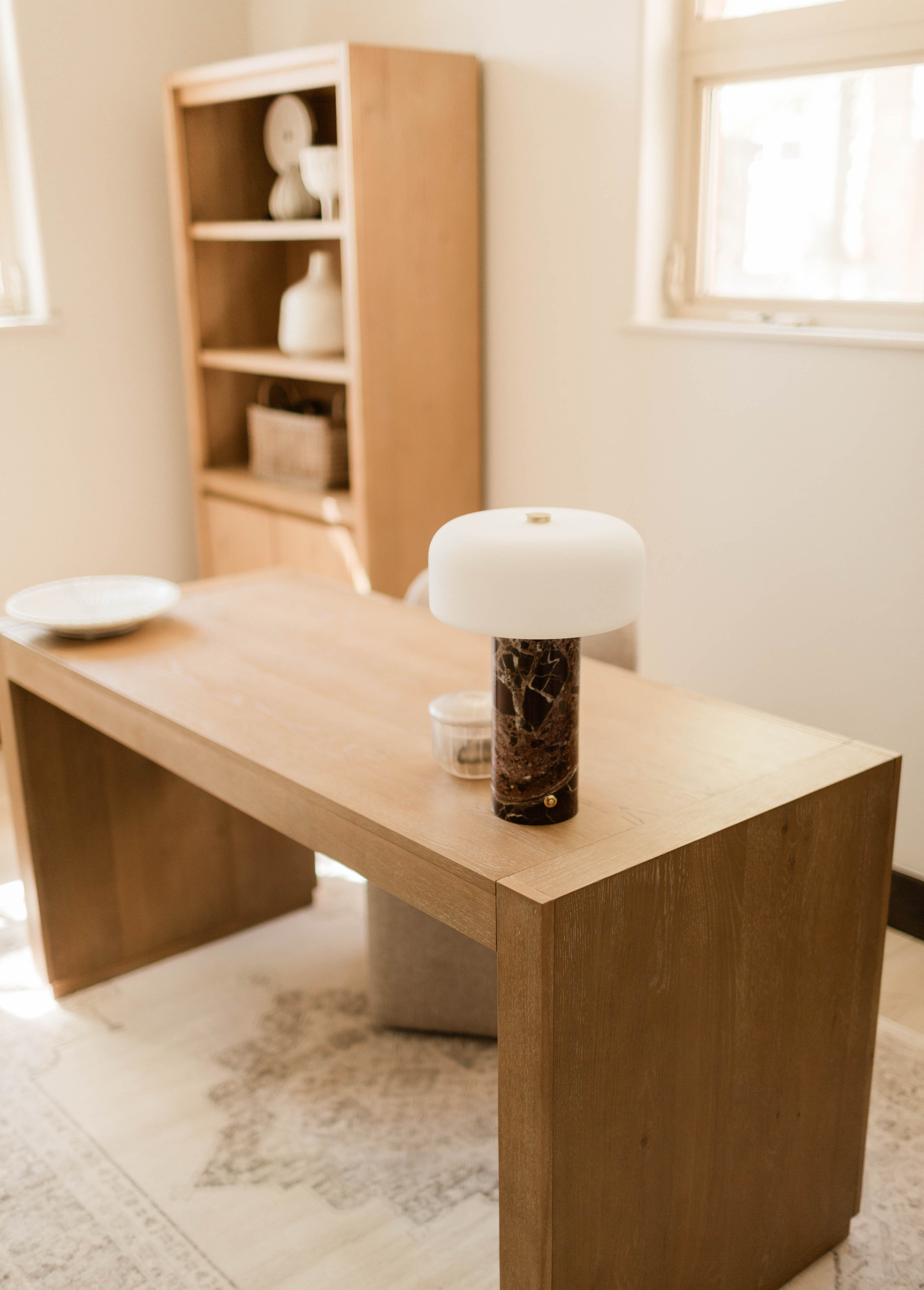 Light wood desk in a softly lit room with a white-topped lamp with a dark marbled base, a white plate, and a small clear container on top, with a wood shelving unit and windows in the background.