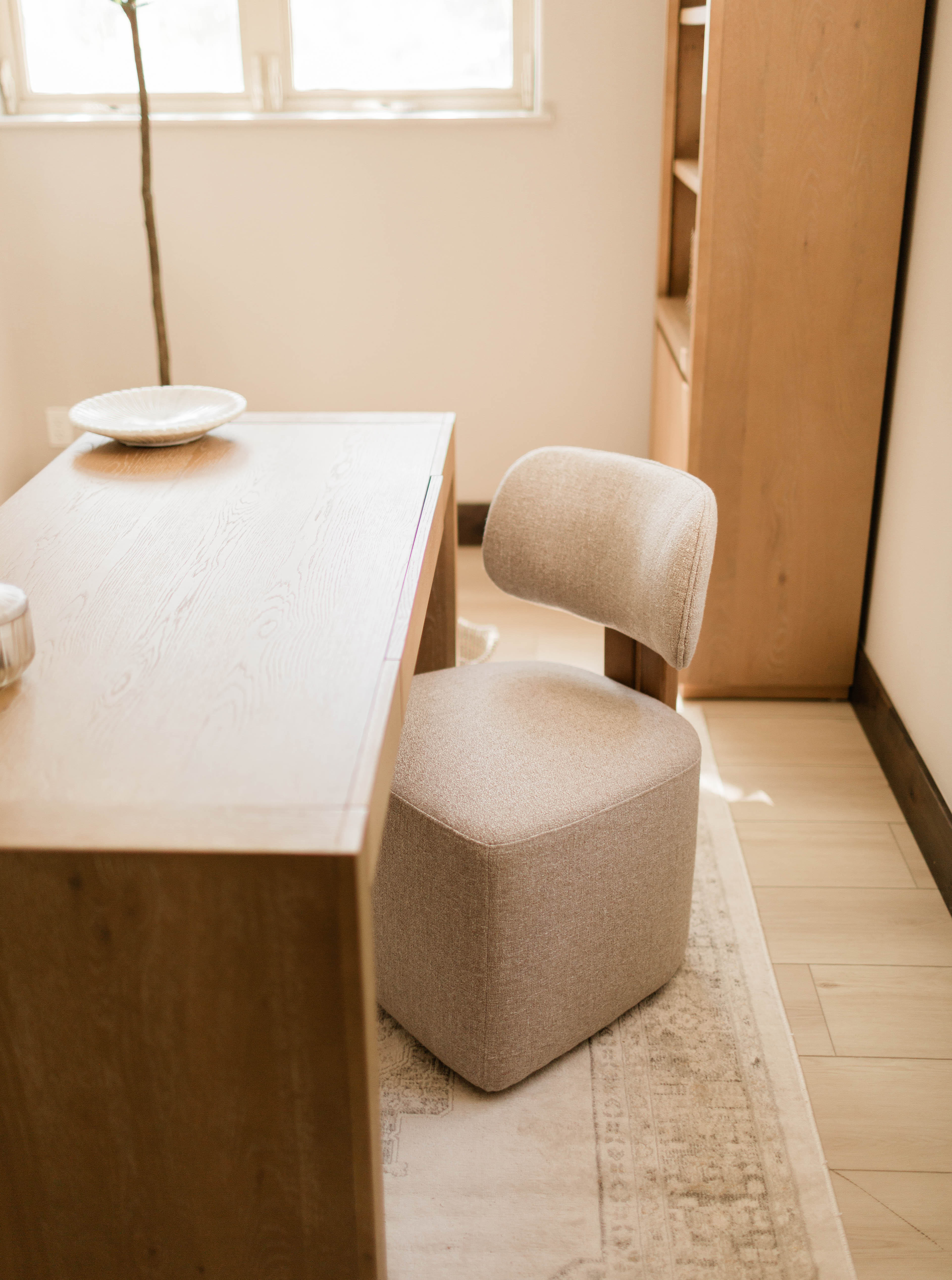 Light beige cushioned chair with rounded backrest next to a wooden desk on a patterned rug in a room with wooden flooring and a window.