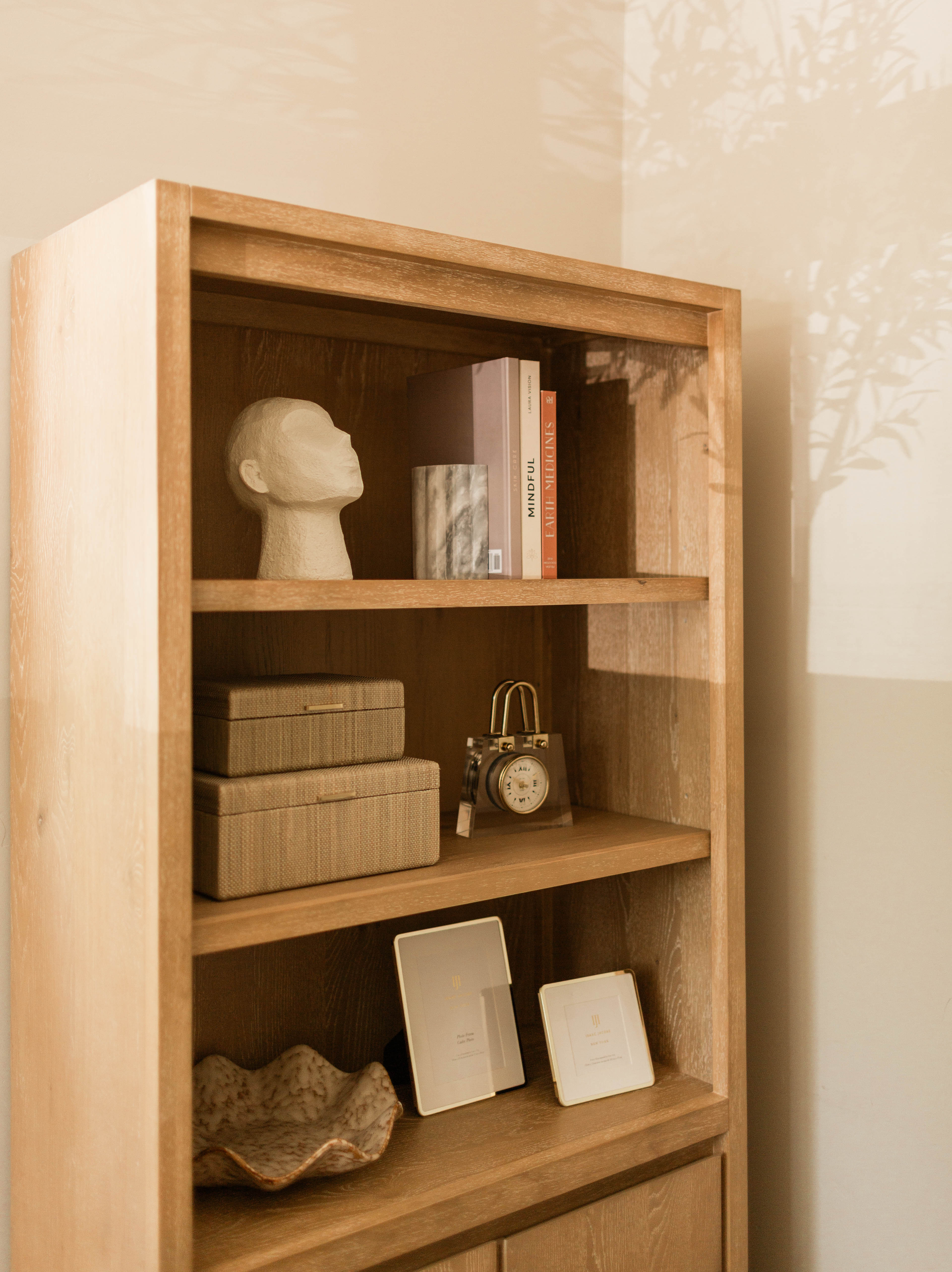 Wooden bookshelf with three shelves holding a sculpted head bust, three books including one titled "MINDFUL", a cylindrical marble container, two stacked woven storage boxes, a small vintage-style clock shaped like a camera, two blank picture frames, and a beige ceramic bowl with a wavy edge, all under soft warm lighting with plant shadows on the wall.
