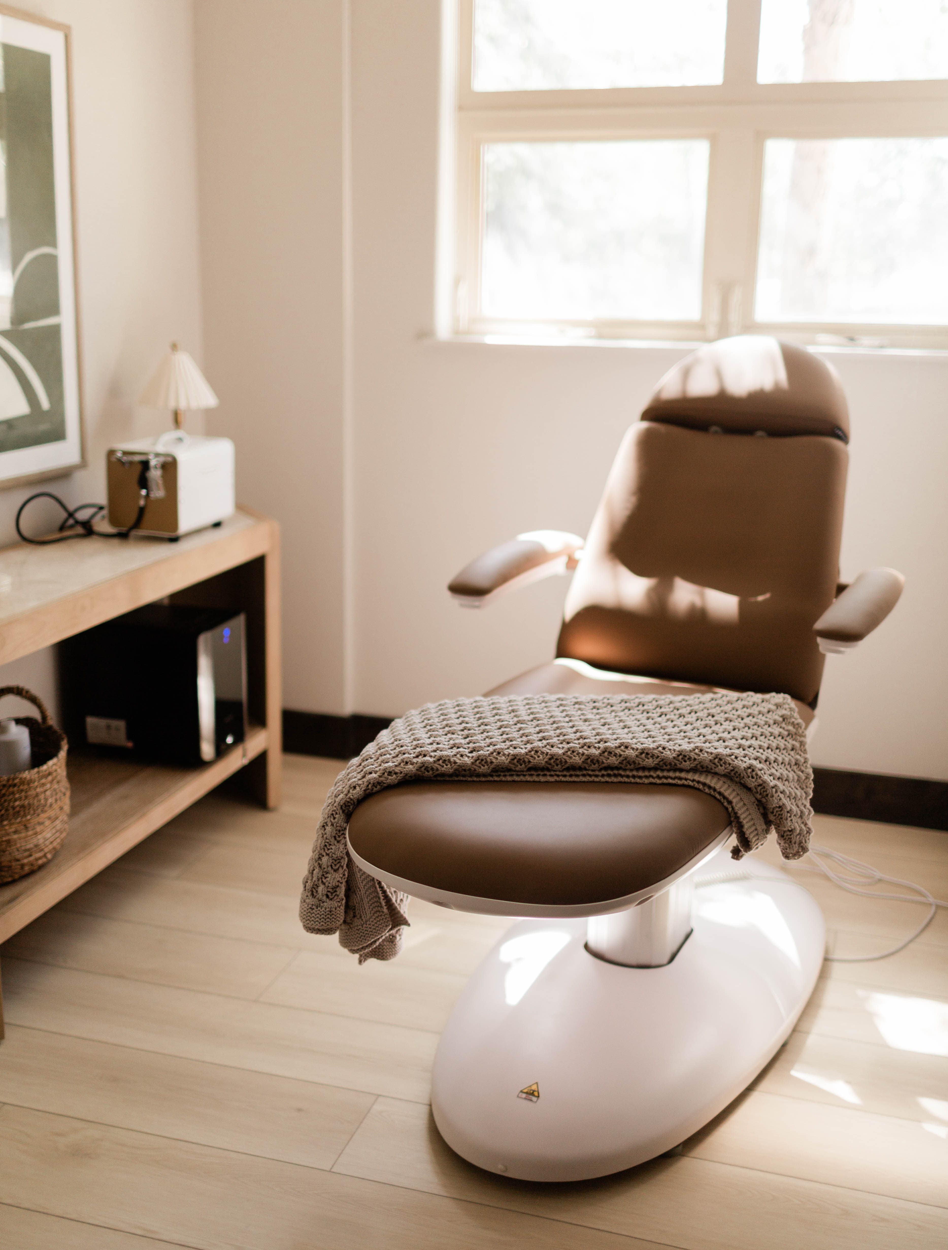 Modern reclining medical or spa chair with a brown cushion and matching armrests, featuring a textured beige blanket draped over the footrest, positioned near a window with soft natural light.