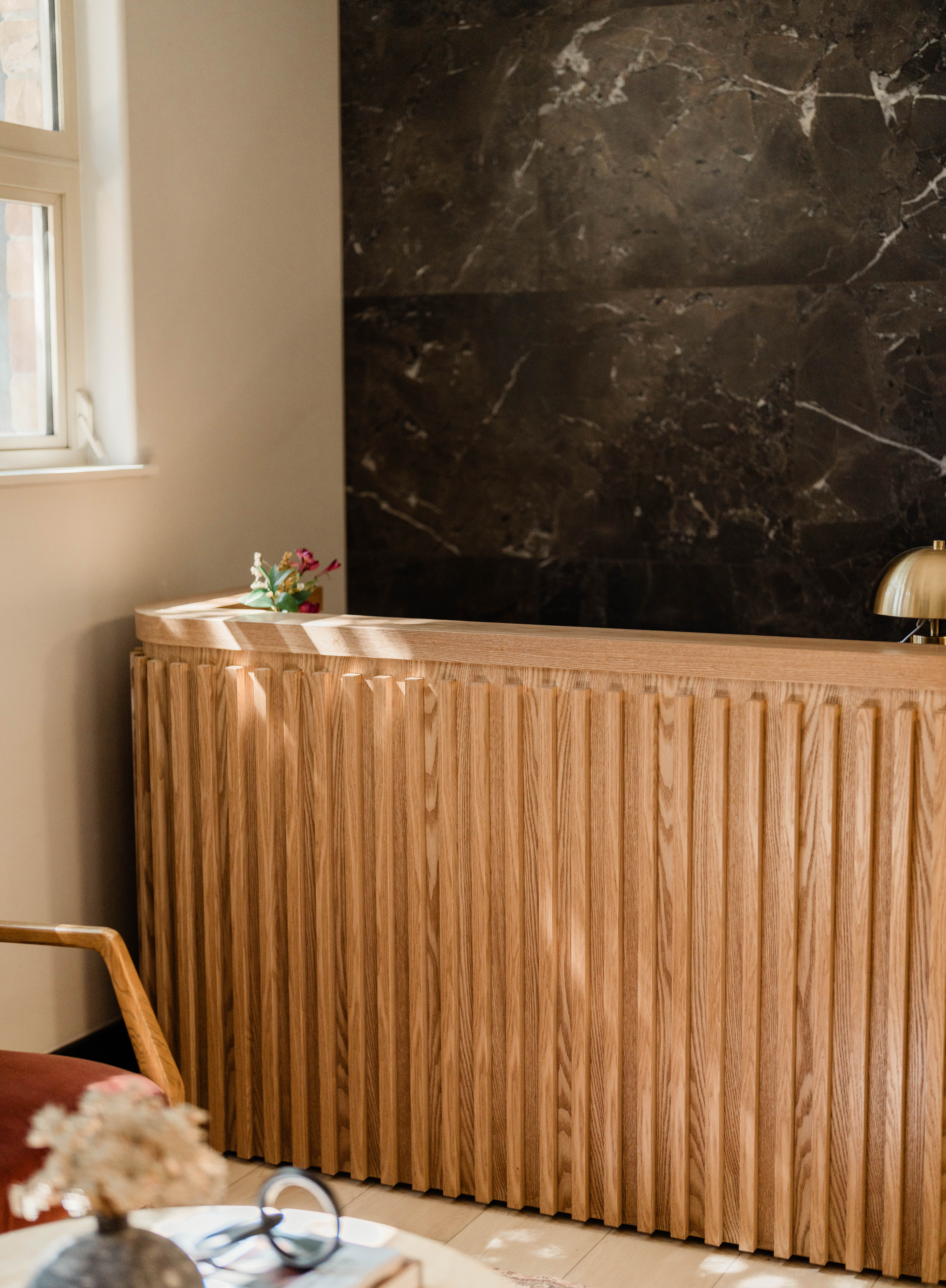 Light wood reception desk with vertical slats in a room with a dark marble wall behind it, a window on the left, a small bouquet of flowers on the desk, and part of a wooden chair and blurred table with eyeglasses and a book in the foreground.