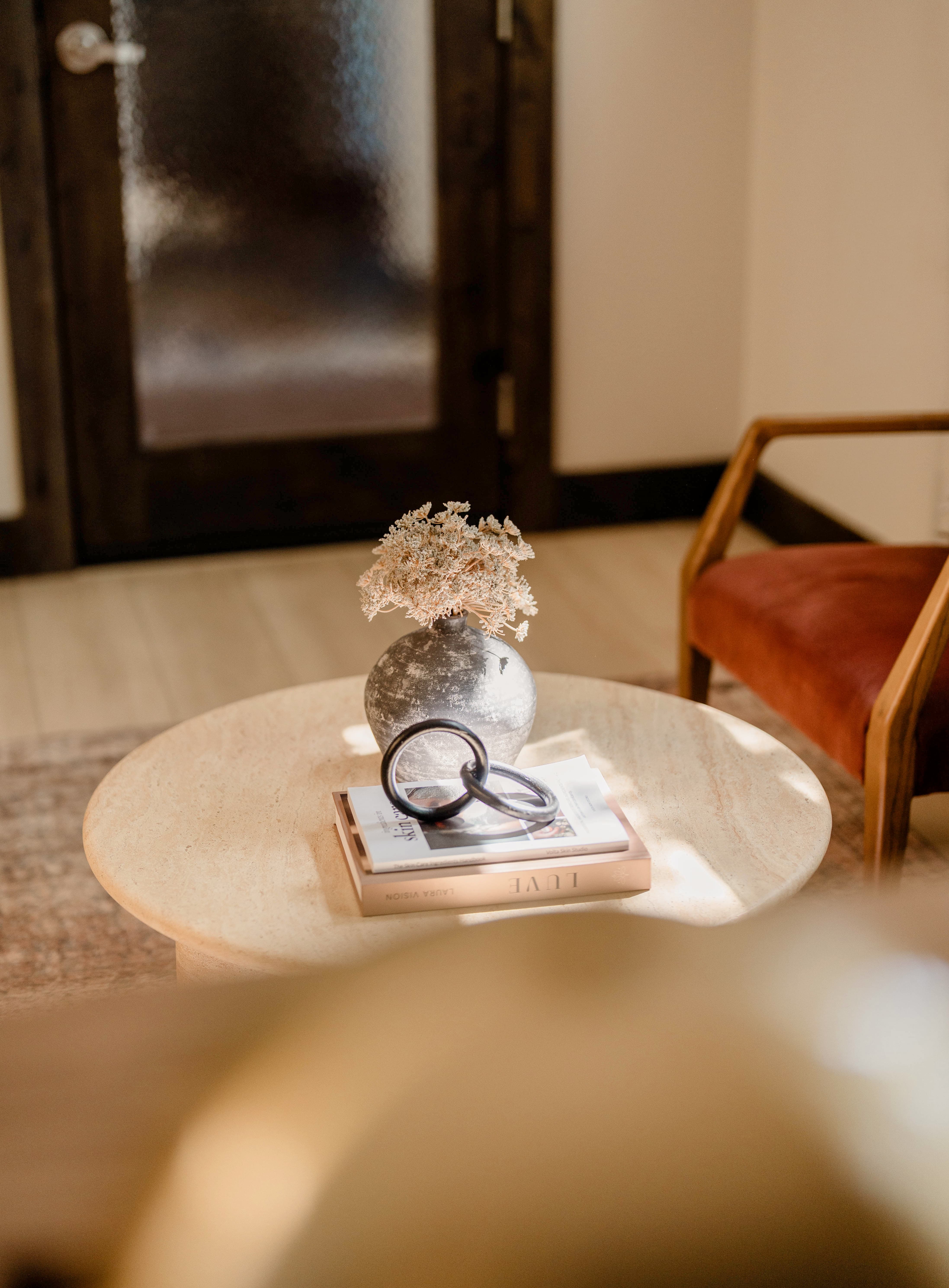 Round marble coffee table with a gray vase holding dried flowers, two stacked books, and a decorative black linked ring ornament, with a brown upholstered chair and wooden-framed frosted glass door in soft natural light.