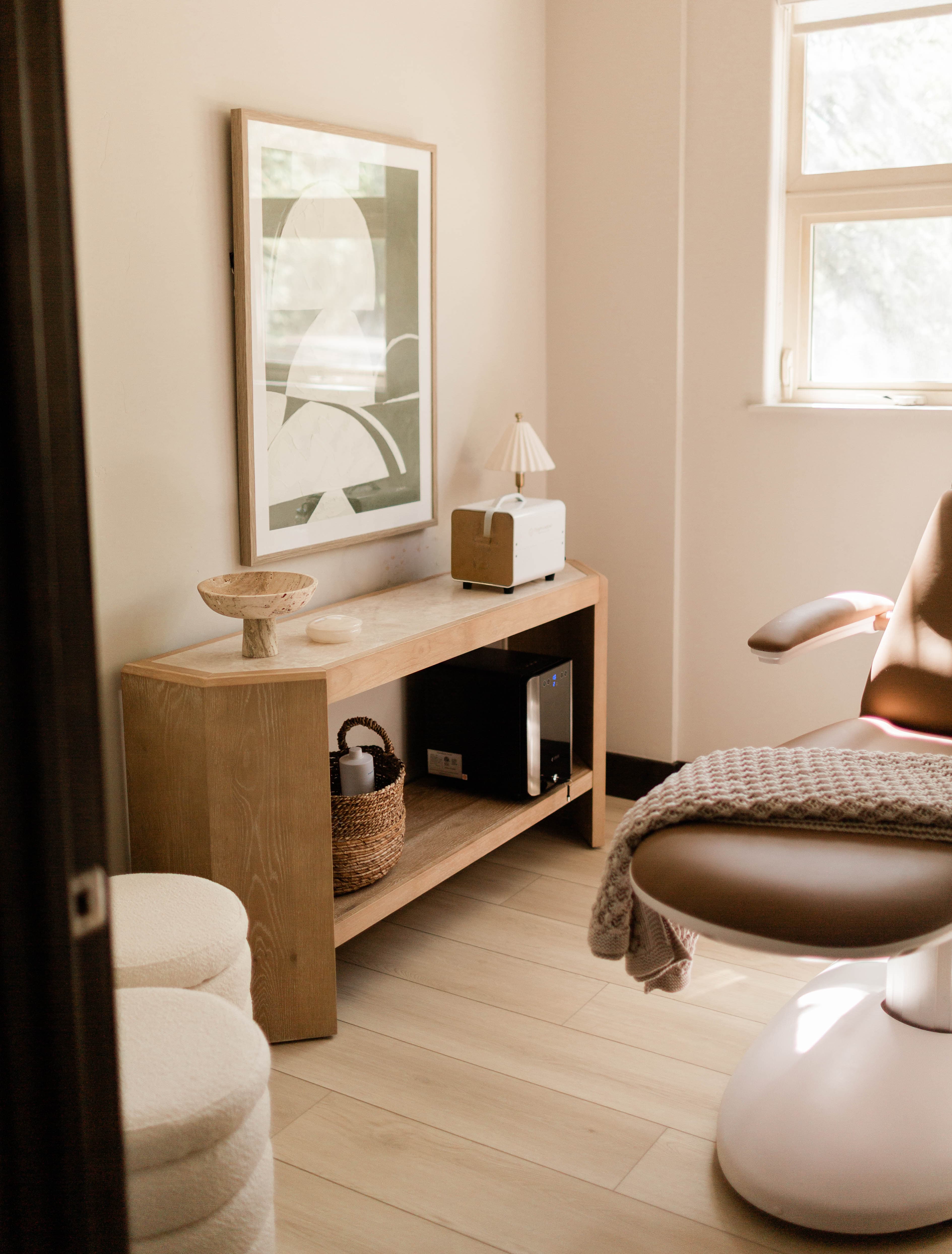 Cozy corner of a room with light wood floor, a wooden console table holding a small lamp, a basket with a bottle, a black mini fridge, and abstract framed artwork on the wall, next to a brown modern chair with a knitted blanket draped over it, under natural light from a window.