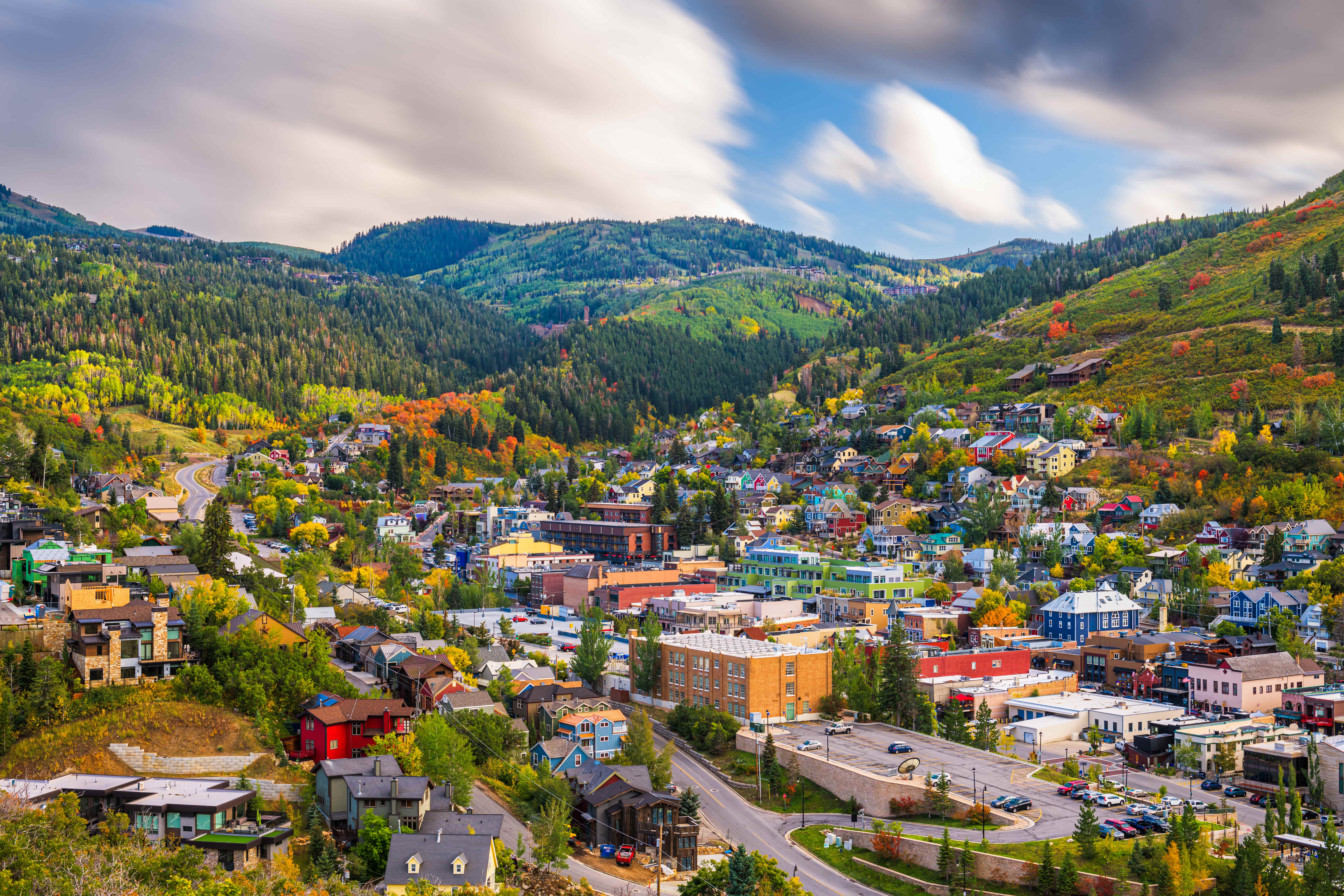 A colorful small mountain town with houses and buildings nestled in a valley surrounded by green and autumn-tinged trees under a partly cloudy sky.