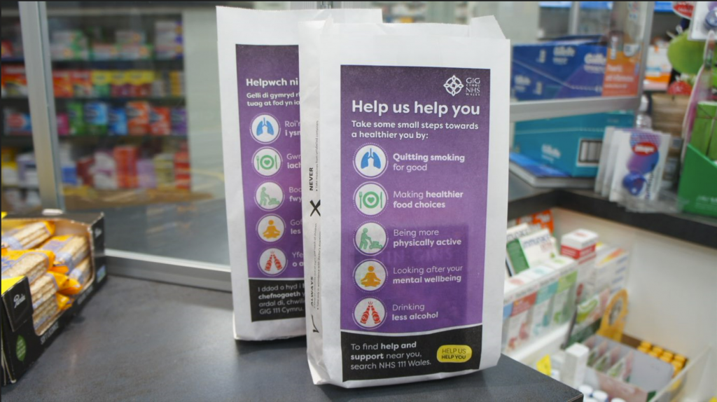 Two purple NHS Wales "Help us help you" pharmacy bags, one in Welsh and one in English, on a pharmacy counter.
