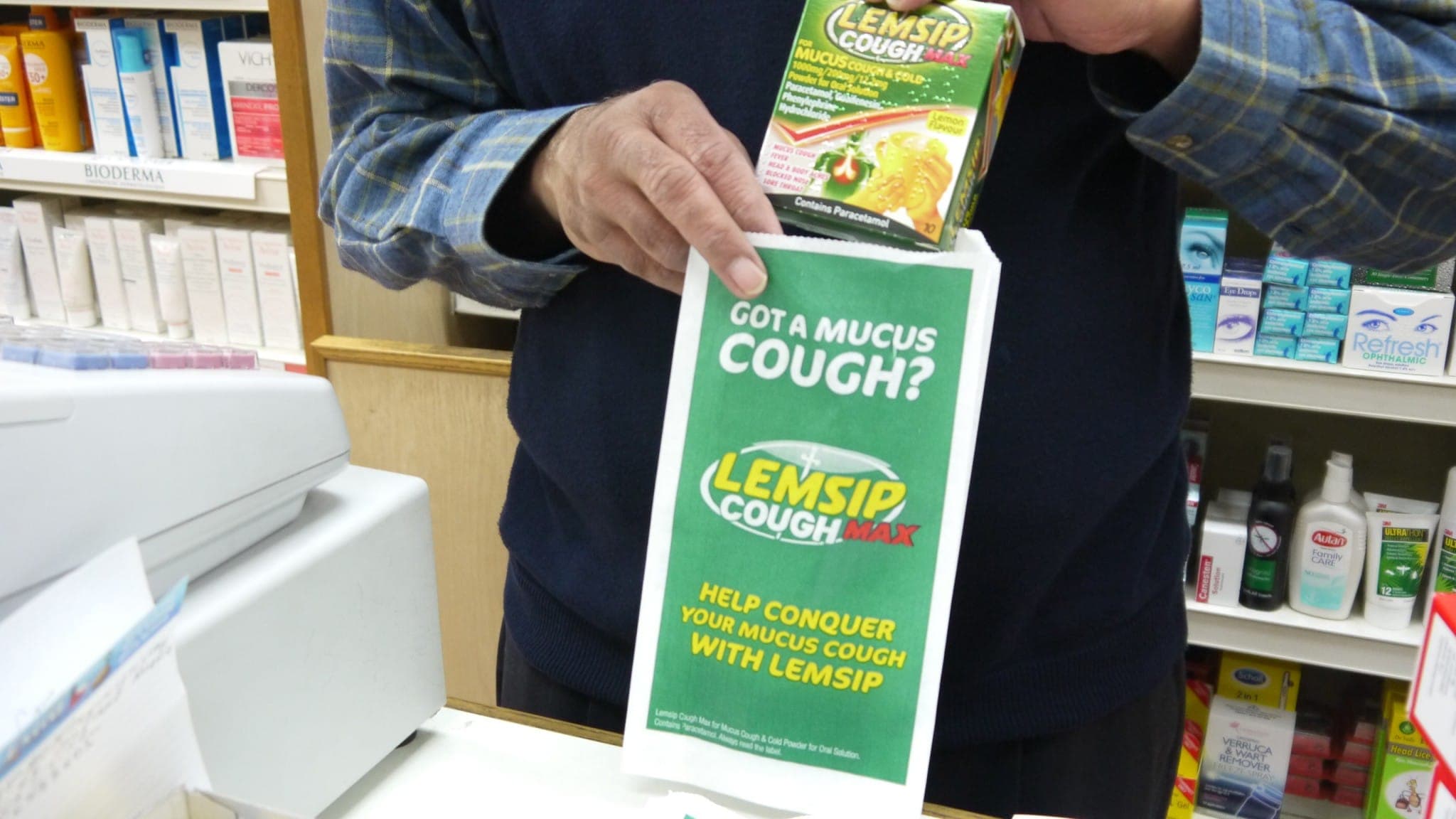 Pharmacist holding a Lemsip Cough Max box above a green Lemsip branded pharmacy bag reading 'Got a mucus cough?'