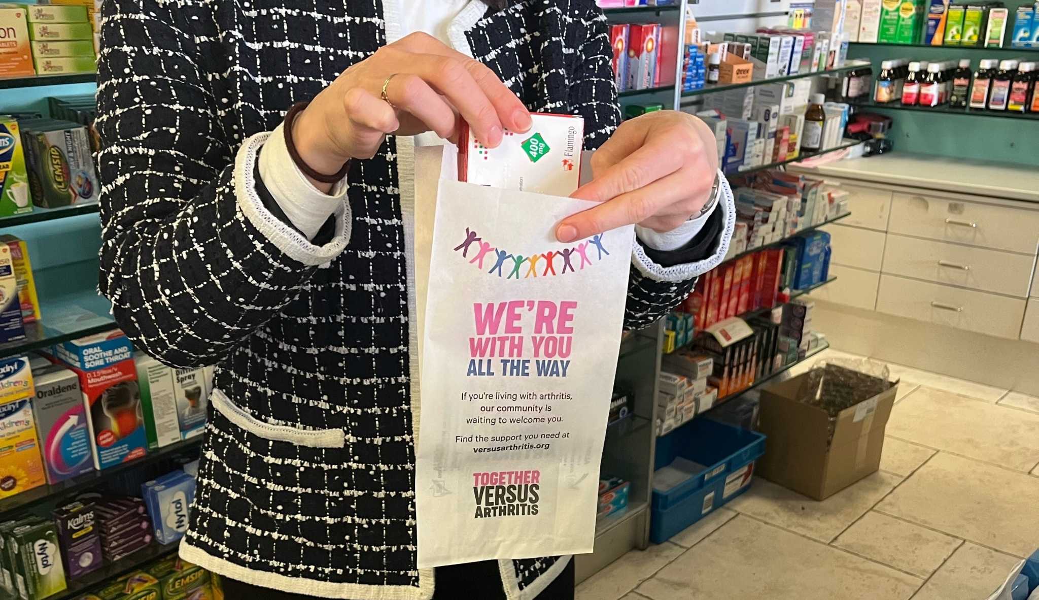 Pharmacist placing medication into a Versus Arthritis pharmacy bag reading 'We're with you all the way' in a pharmacy