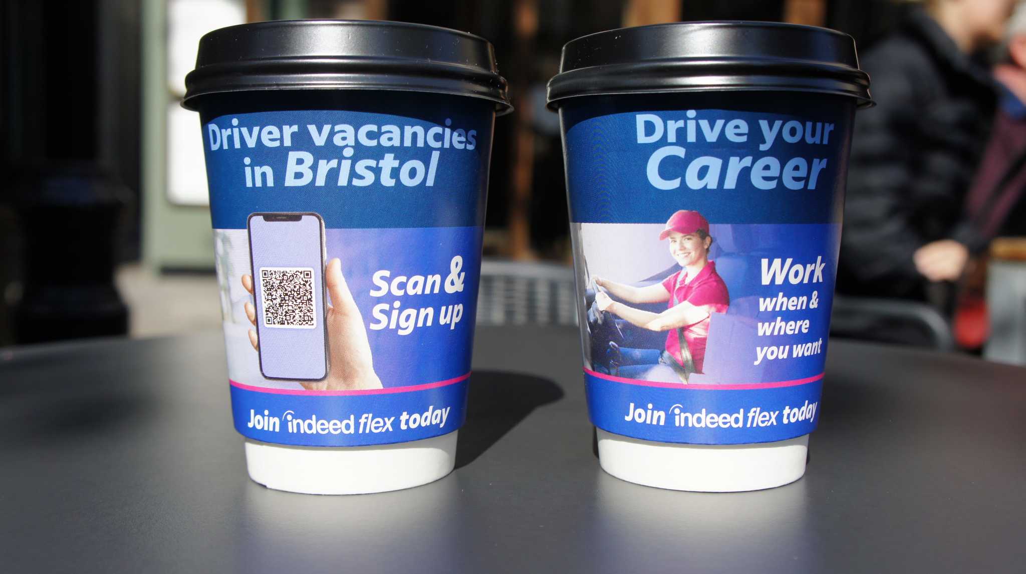 Two blue Indeed Flex branded coffee cups advertising driver vacancies in Bristol with a QR code, on an outdoor table