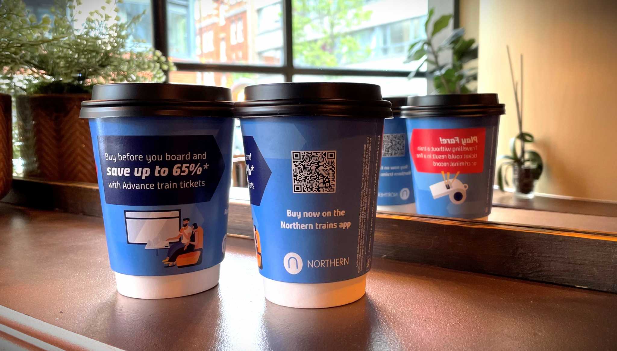 Blue Northern Rail branded coffee cups promoting advance train ticket savings with a QR code for the Northern app, on a windowsill