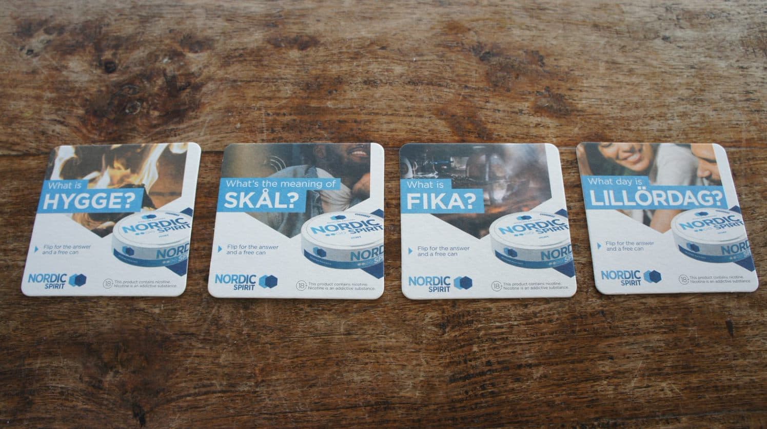 Four Nordic Spirit branded beer mats asking about Scandinavian words Hygge, Skal, Fika and Lillordag, laid on a wooden table
