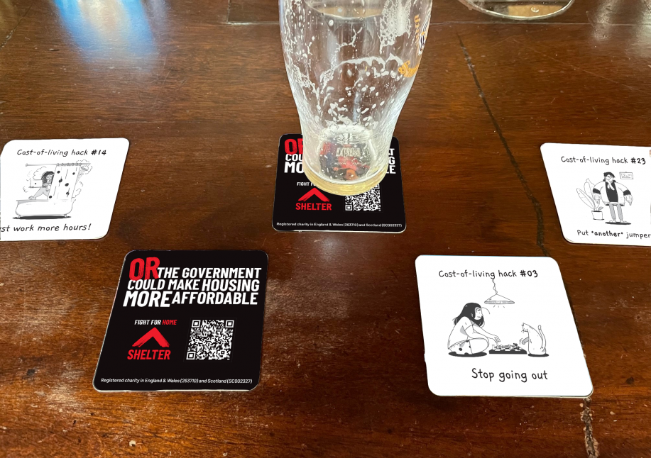 Shelter beer mats on a pub table, with cost-of-living hack cartoons and the reverse reading 'Or the government could make housing more affordable'