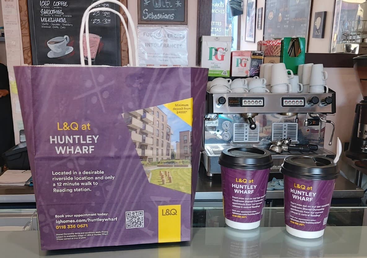 Purple L&Q Huntley Wharf branded deli bag and two coffee cups on a cafe counter in front of a coffee machine