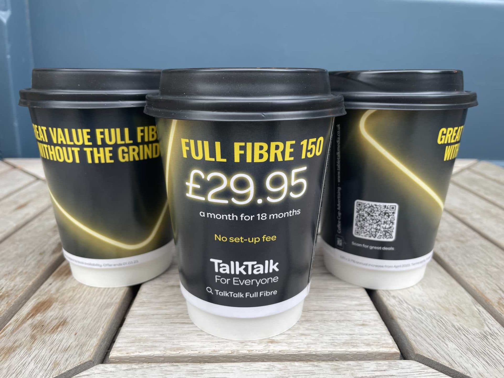Three black TalkTalk branded coffee cups advertising Full Fibre 150 broadband for £29.95 a month, on a wooden table