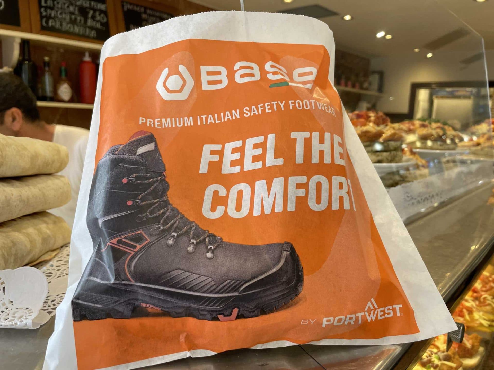 Orange Base safety footwear by Portwest bacon butty bag reading 'Feel the comfort' with a work boot, on a sandwich shop counter