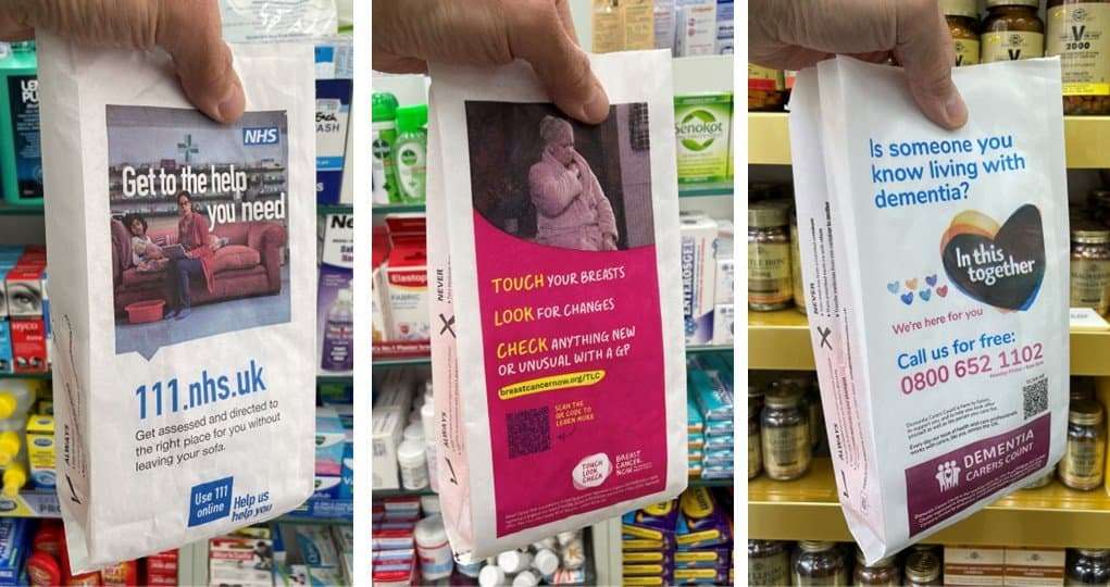 Three branded pharmacy bags held up in a pharmacy, advertising NHS 111, Breast Cancer Now Touch Look Check and Dementia Carers Count
