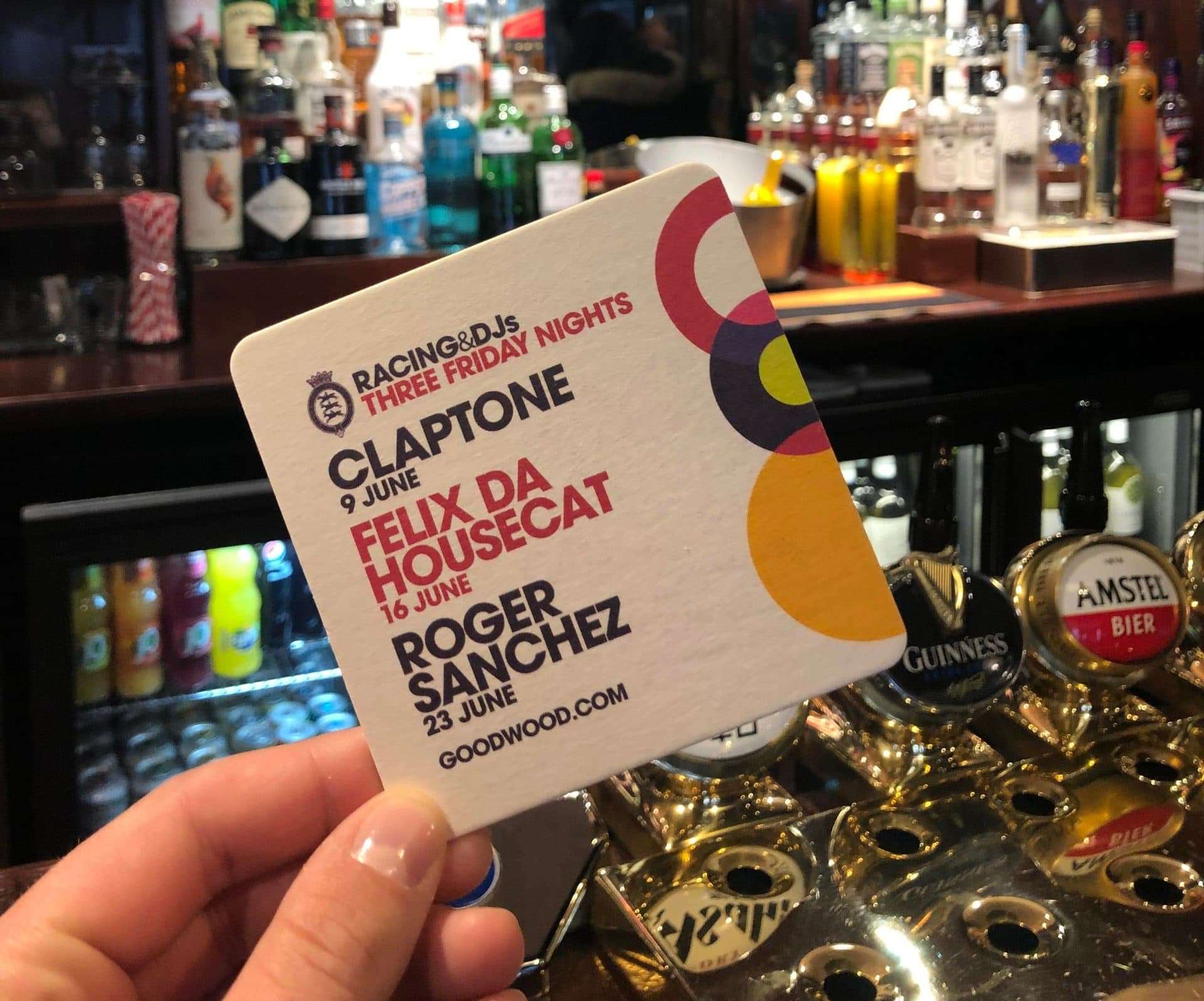 Hand holding a Goodwood Racing & DJs Three Friday Nights beer mat listing Claptone, Felix Da Housecat and Roger Sanchez, in front of a pub bar