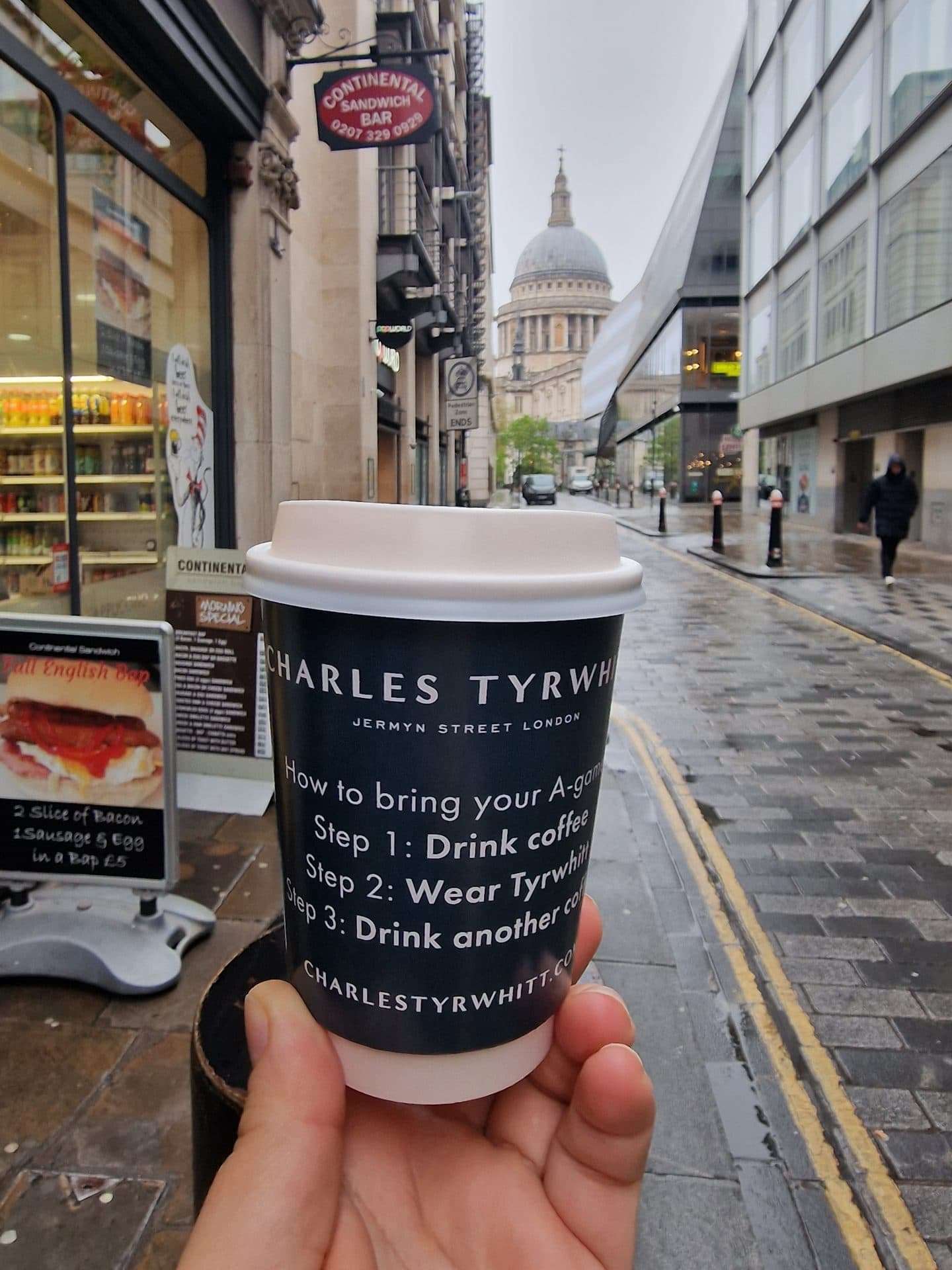 Hand holding a black Charles Tyrwhitt branded coffee cup on a London street with St Paul's Cathedral in the background