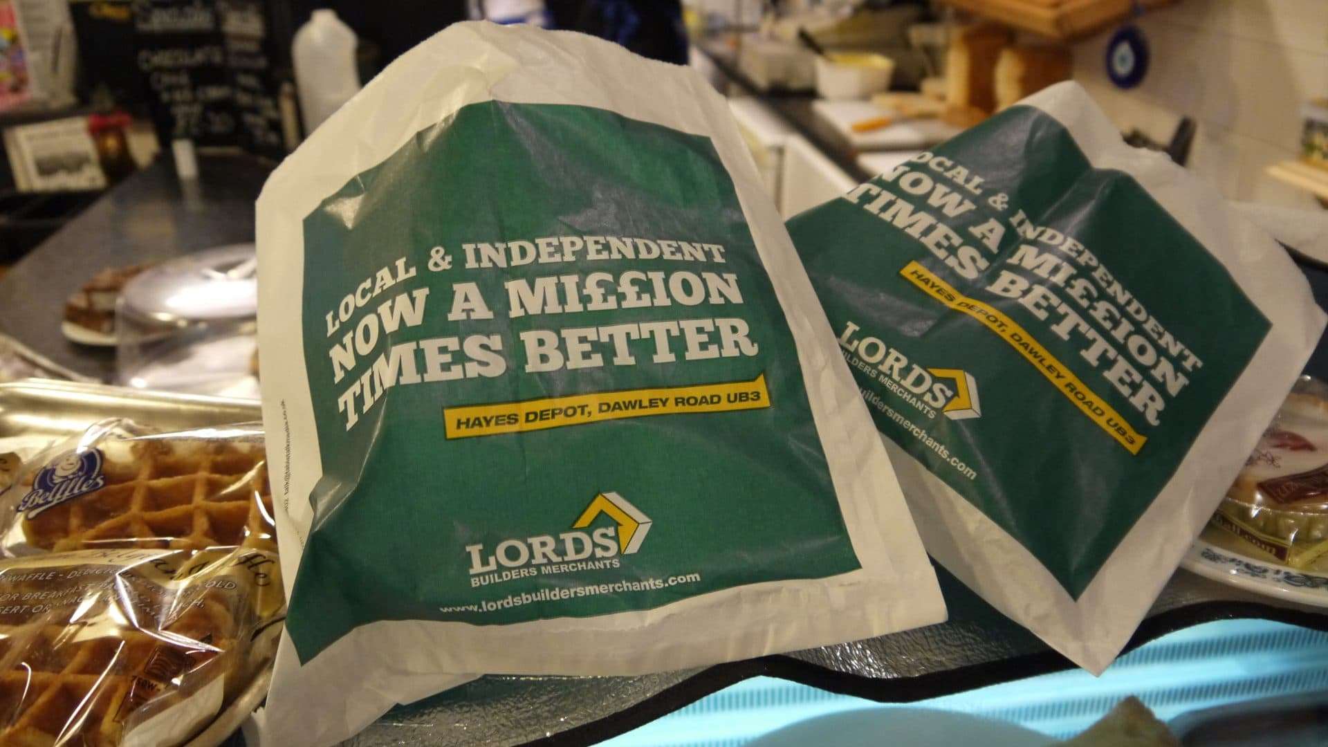 Two green Lords Builders Merchants sandwich bags reading 'Local & independent, now a million times better' for the Hayes depot, on a cafe counter