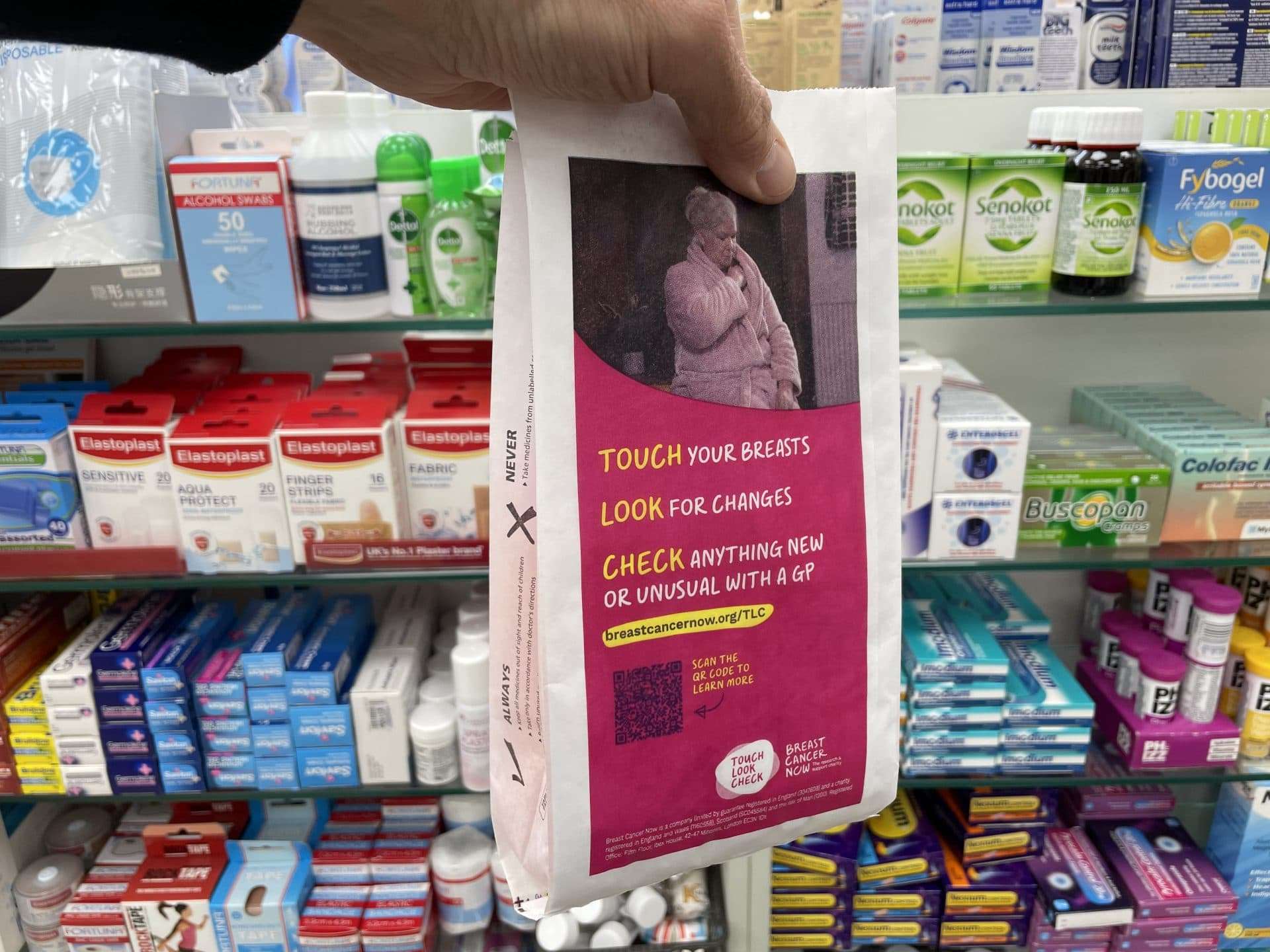 Hand holding a pink Breast Cancer Now pharmacy bag with the Touch, Look, Check message and a QR code, in front of pharmacy shelves