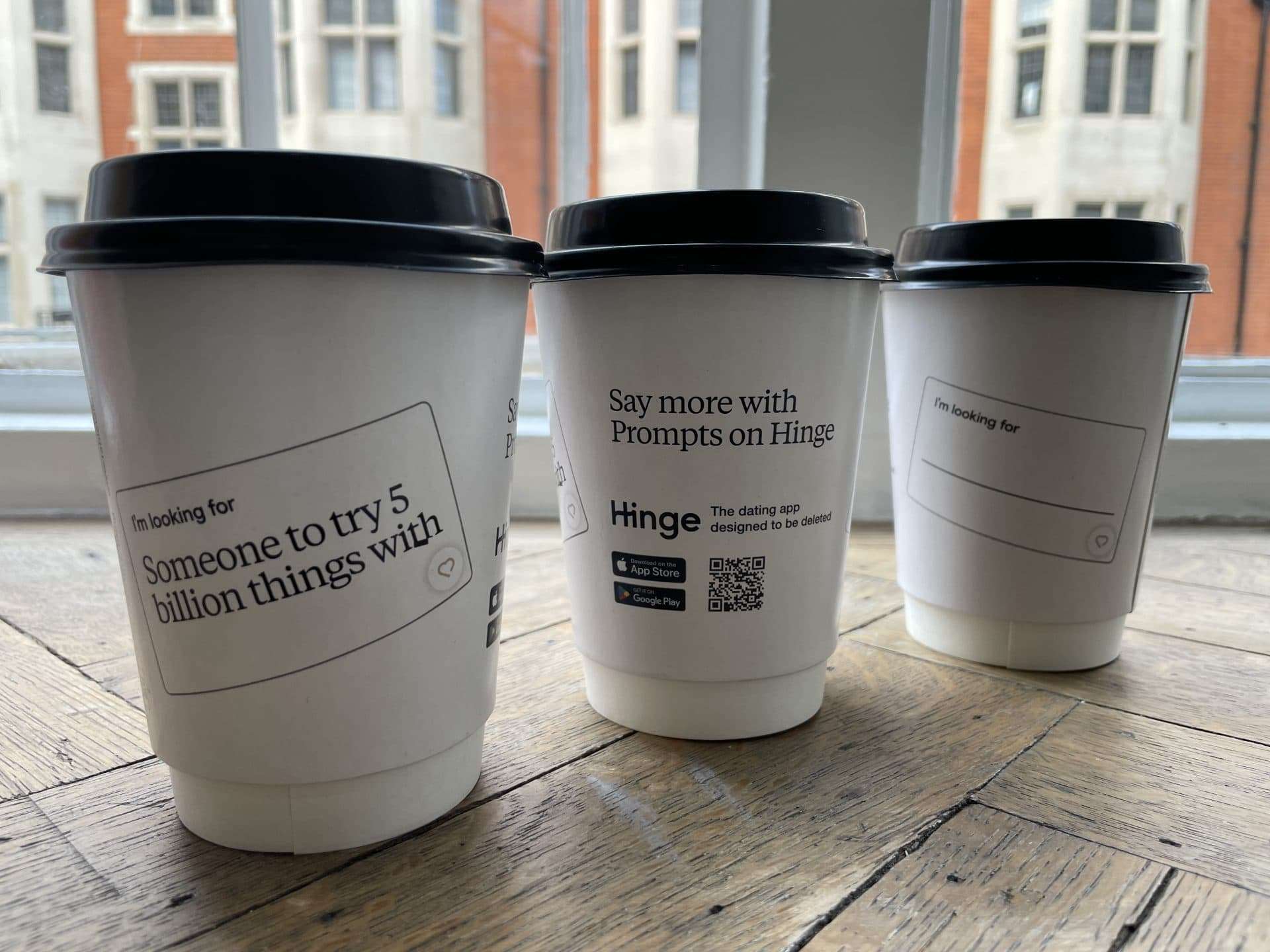 Three white Hinge dating app takeaway coffee cups with 'I'm looking for' prompts and a QR code, on a wooden floor by a window