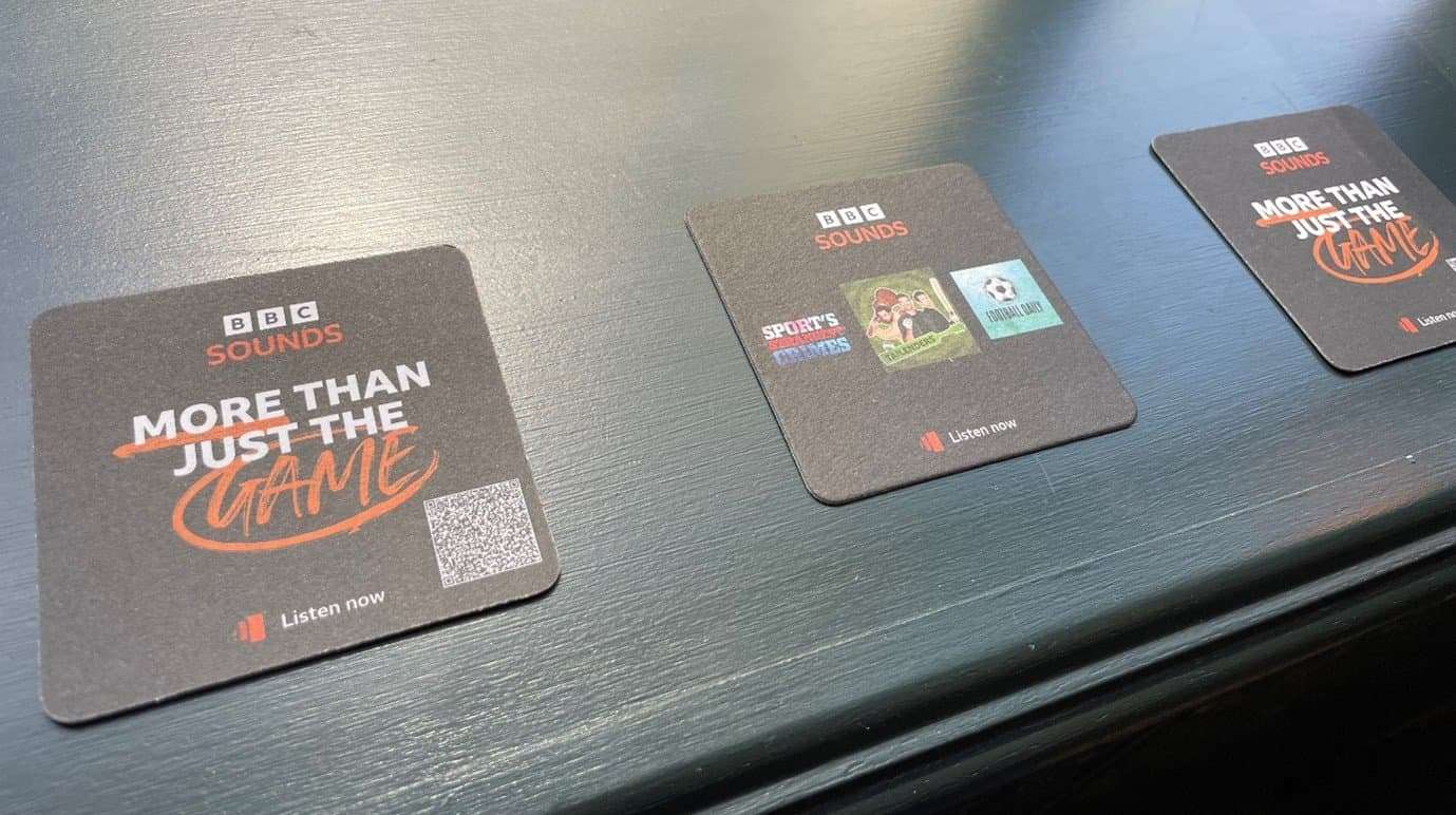 Black BBC Sounds beer mats reading 'More than just the game' with a QR code and sports podcast artwork, on a dark pub table