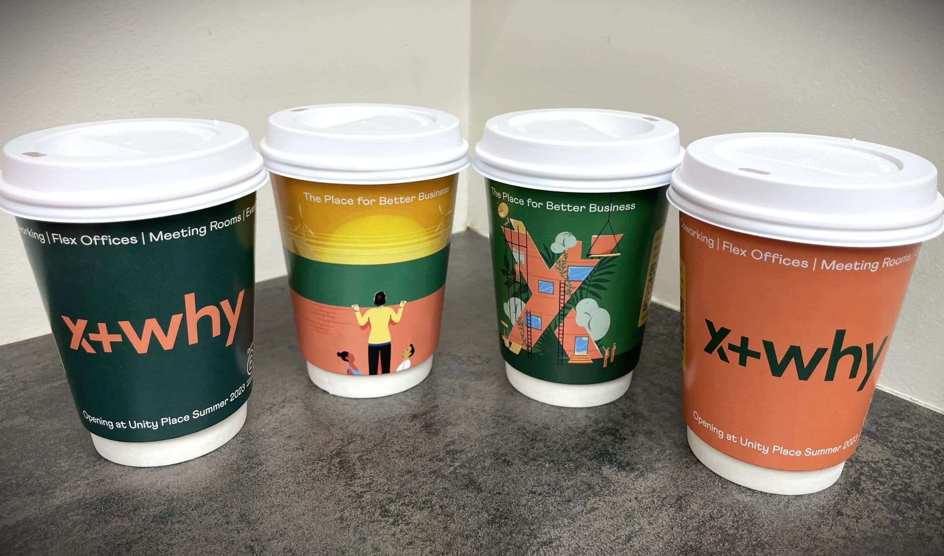 Four green and orange x+why branded coffee cups promoting flex offices and meeting rooms opening at Unity Place, on a grey worktop