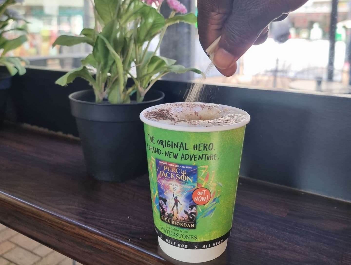 Hand pouring sugar into a green coffee cup advertising the new Percy Jackson novel by Rick Riordan, available from Waterstones