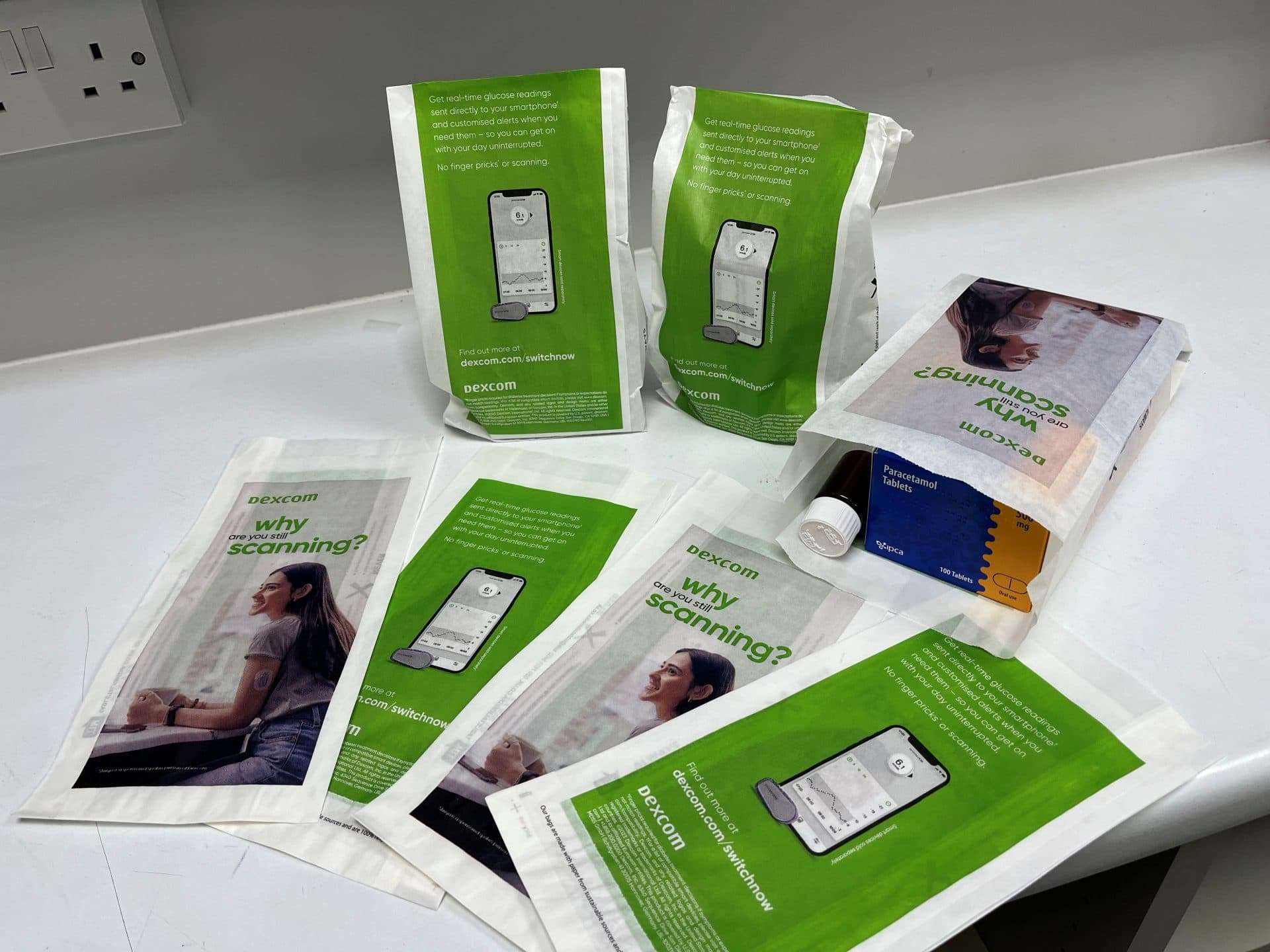 Green Dexcom pharmacy bags asking 'Why are you still scanning?' and showing a glucose monitoring app, laid out on a white counter
