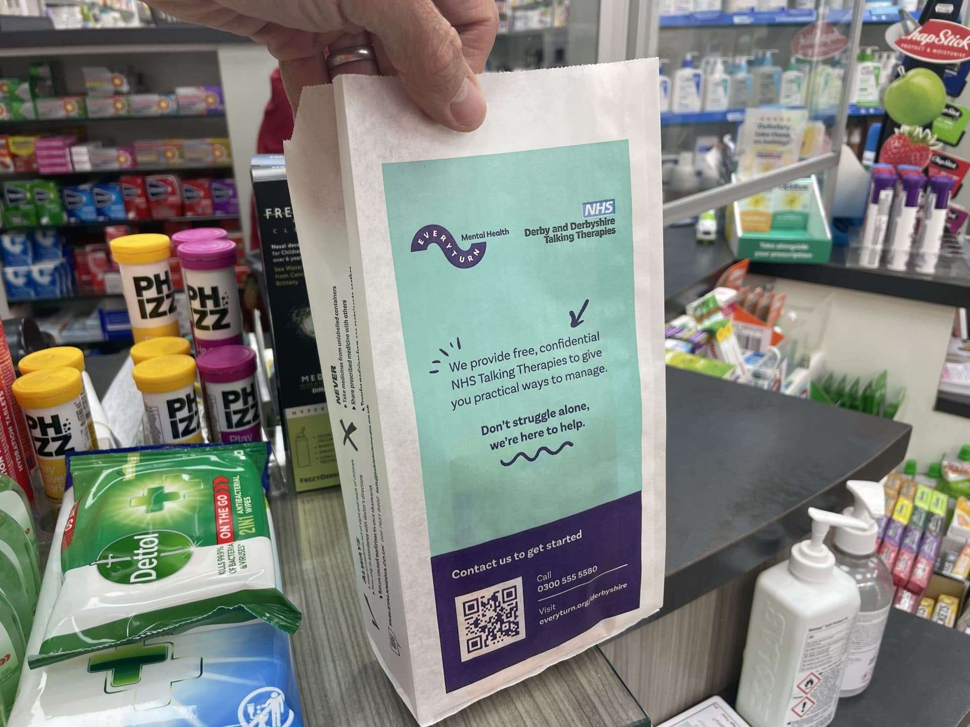 Hand holding an Everyturn Mental Health pharmacy bag promoting free NHS Derby and Derbyshire Talking Therapies, at a pharmacy counter