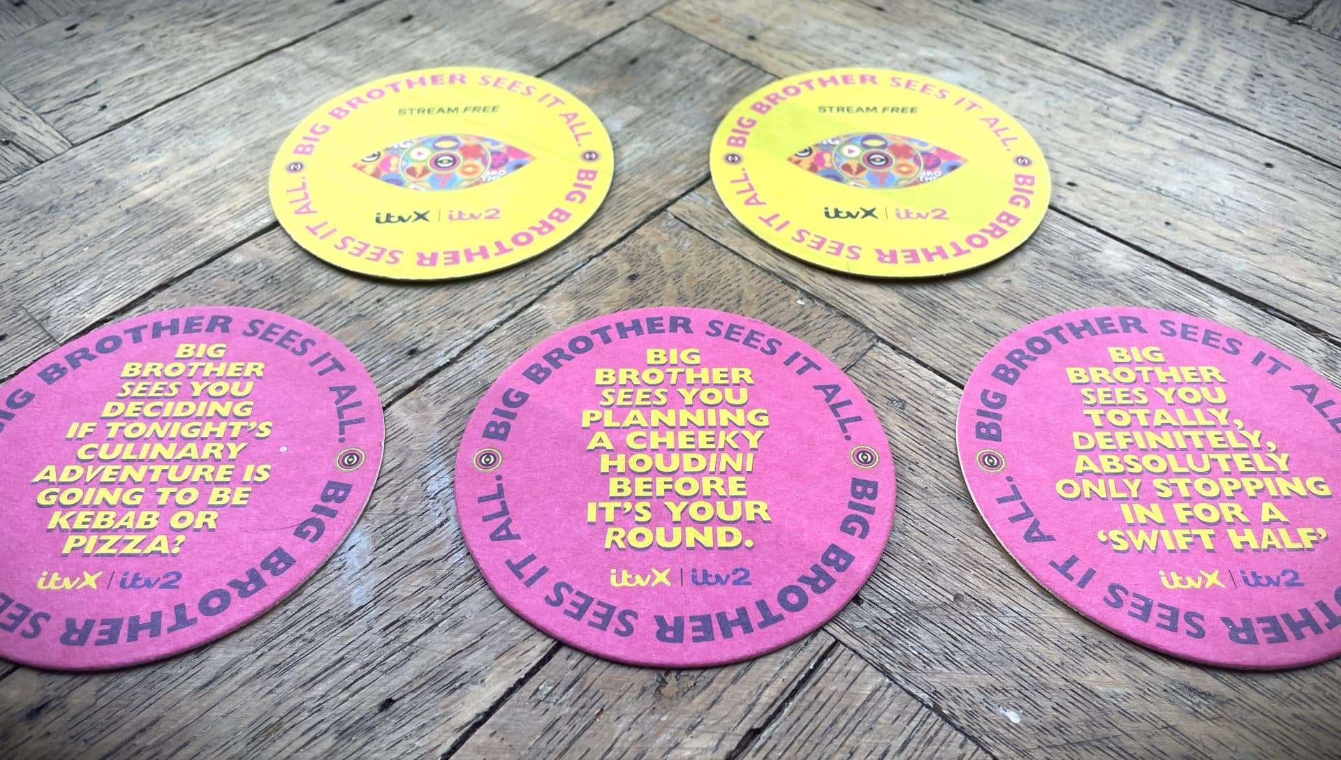 Round pink and yellow ITVX Big Brother beer mats with 'Big Brother sees it all' pub-themed jokes, on a wooden floor