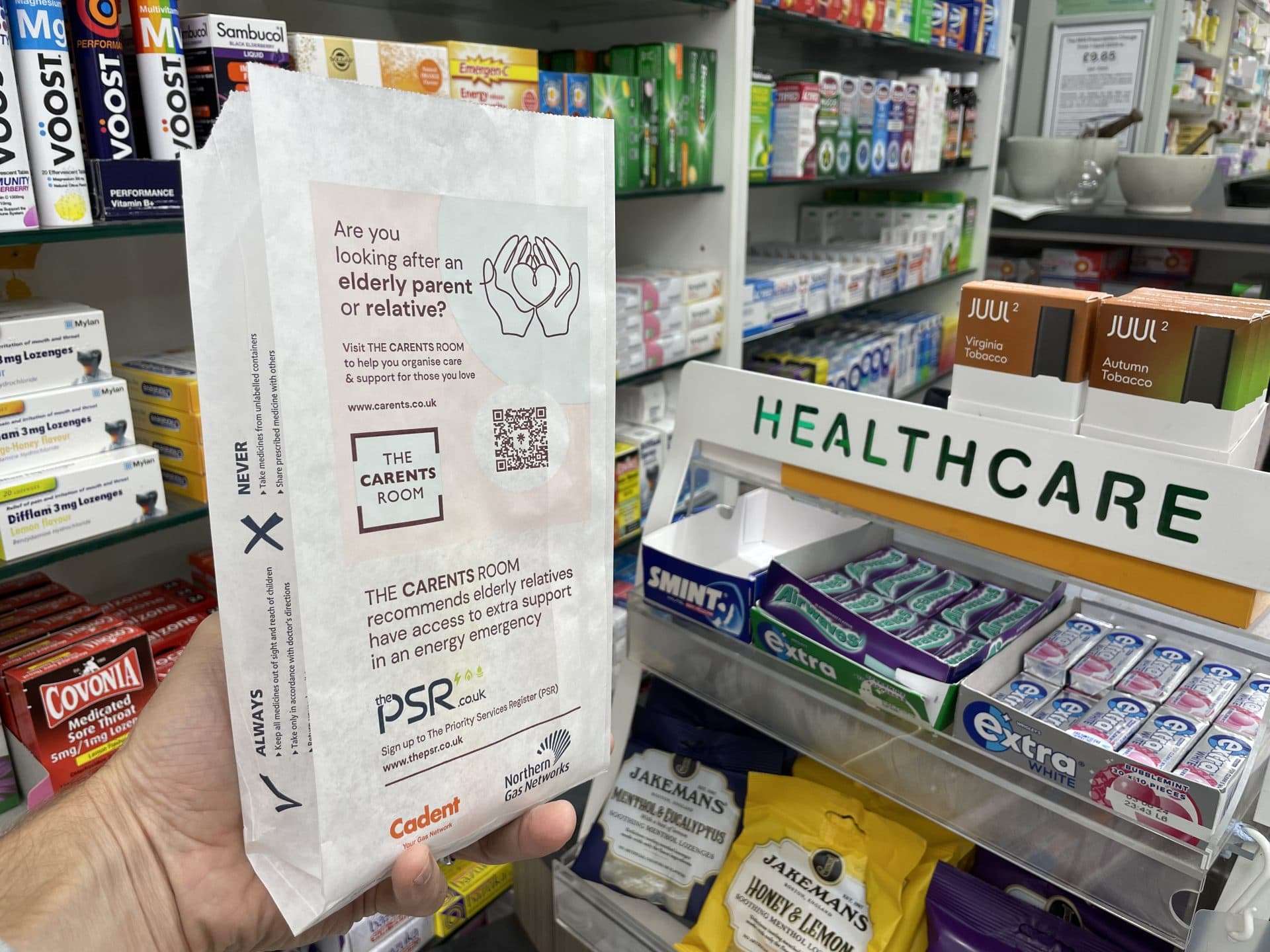 Hand holding a Carents Room pharmacy bag for people caring for elderly relatives, promoting the Priority Services Register, in a pharmacy