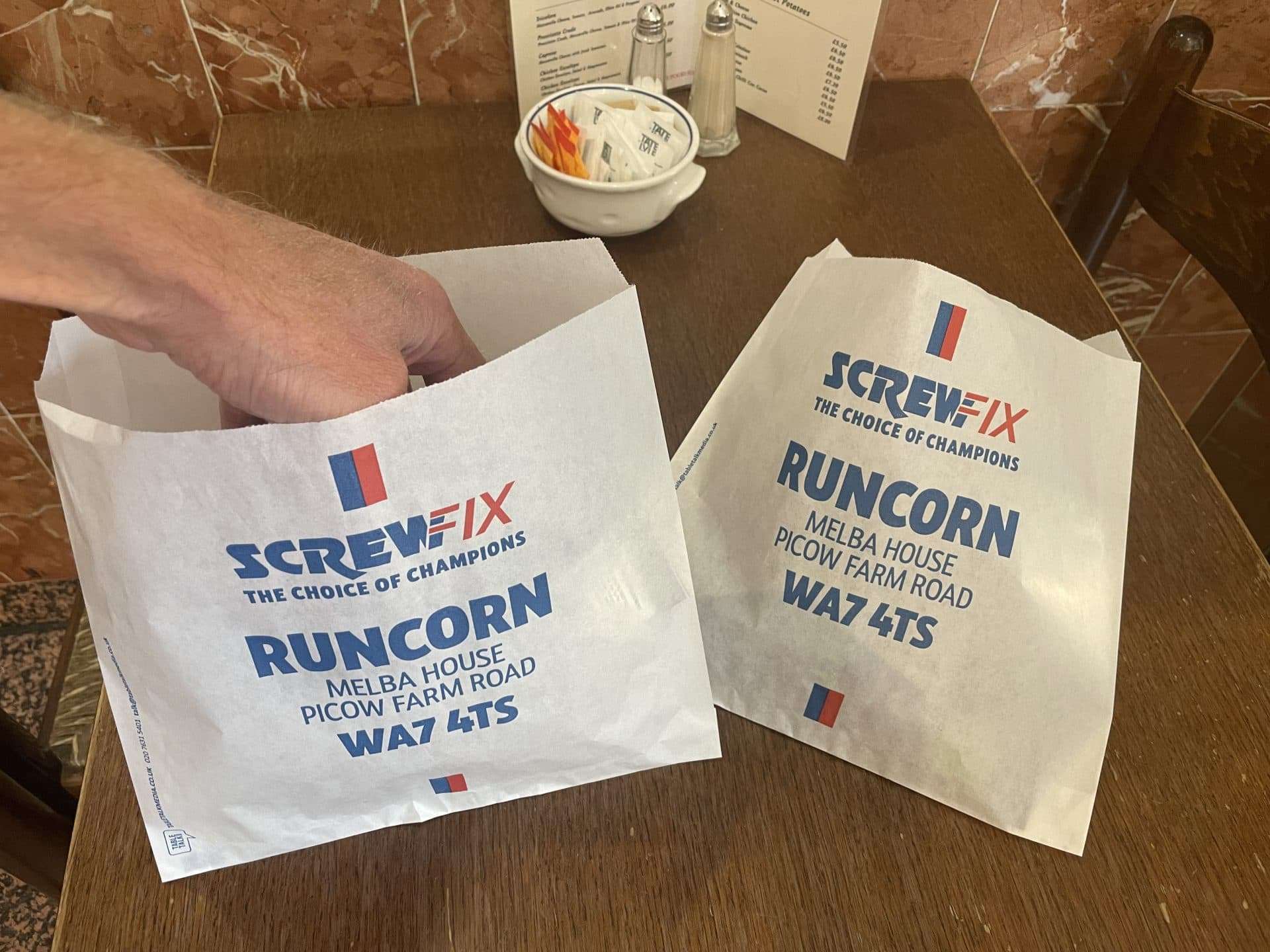 Two white Screwfix bacon butty bags promoting the new Runcorn branch on Picow Farm Road, on a cafe table